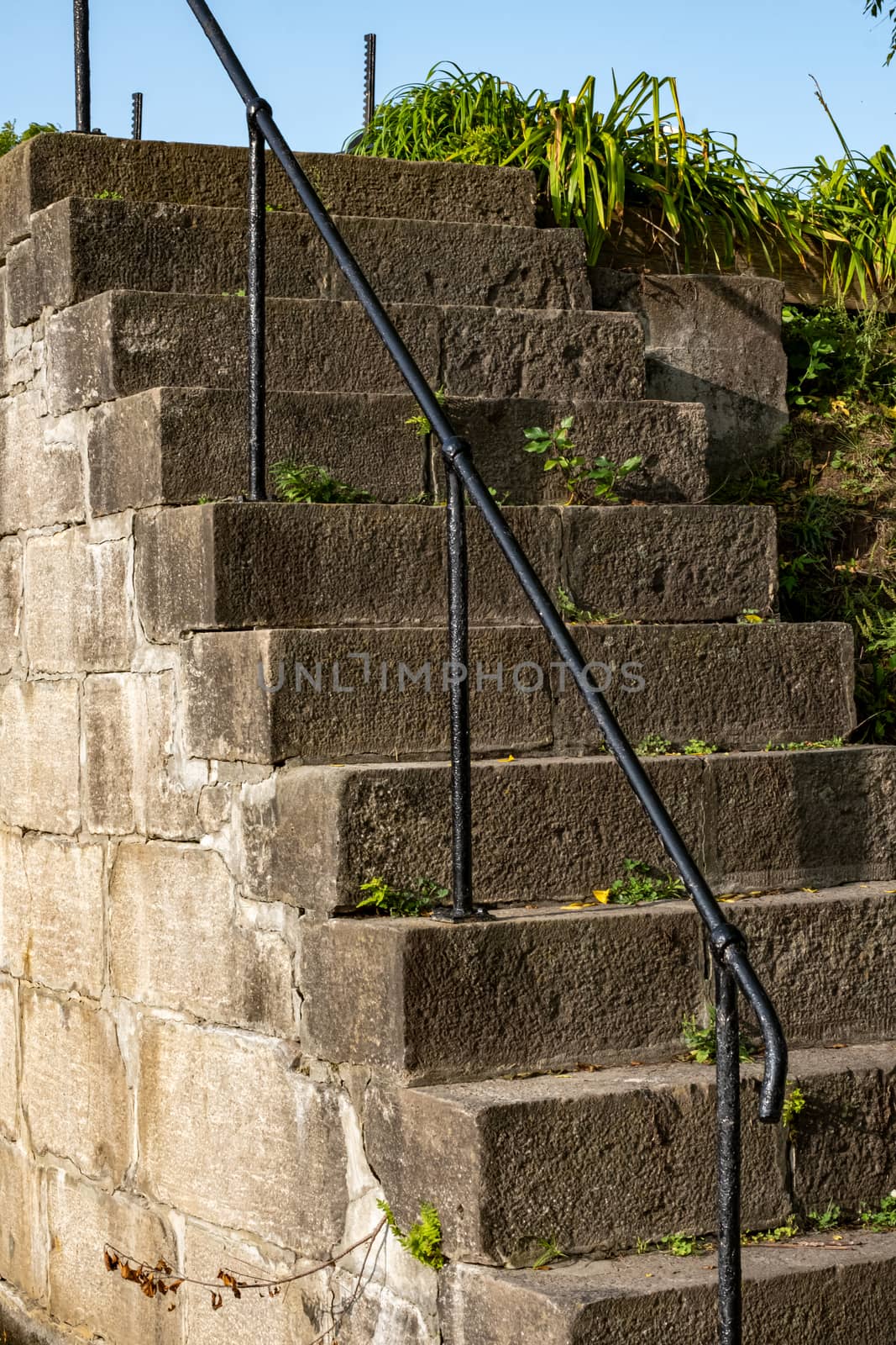 An old stone staircase with an iron railing is embedded into the side of a hill, with plants visible beside it, and growing through its cracks.