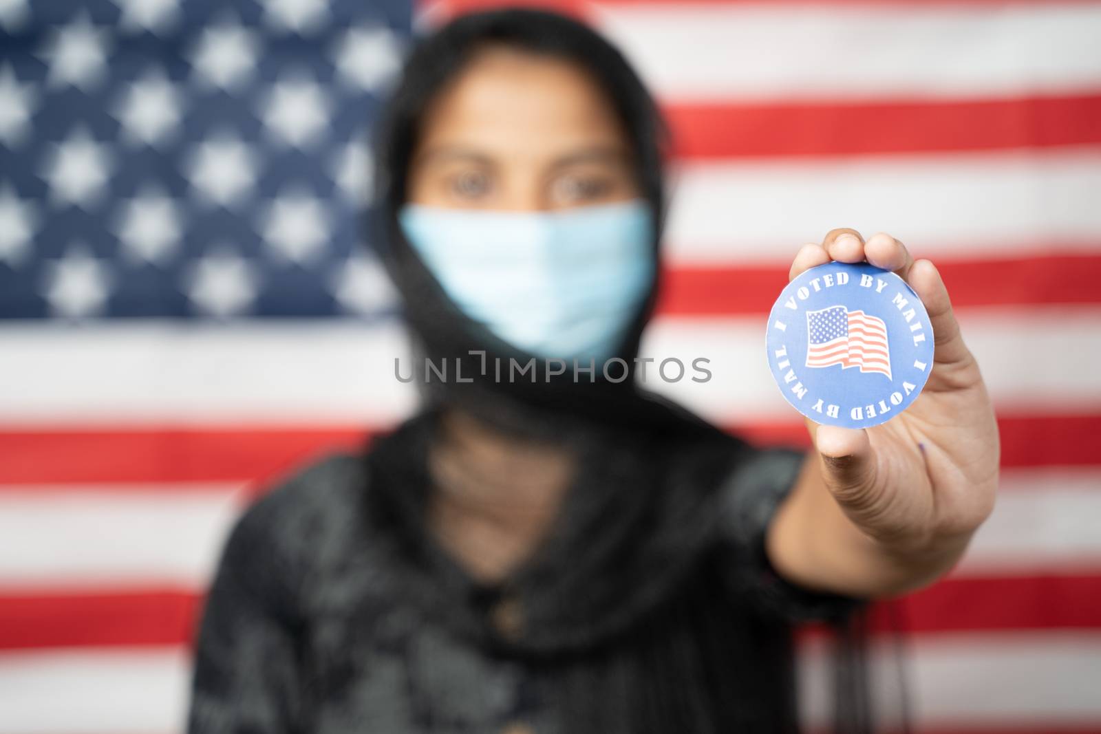 Selective focus on hands, Girl with Hijab or head covering and mask worn showing I voted Sticker with US flag as background - Concept of voting during US election