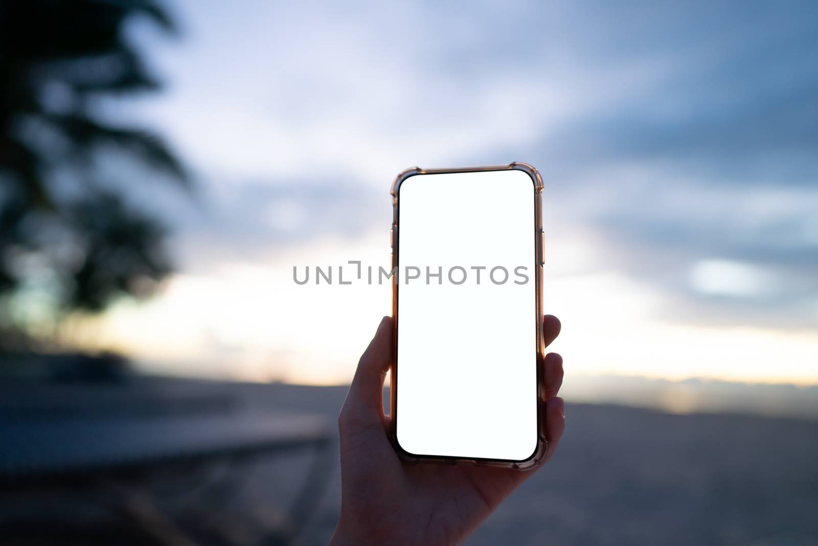Woman hand holding mobile phone with blank white screen mockup on the beach in the morning.
