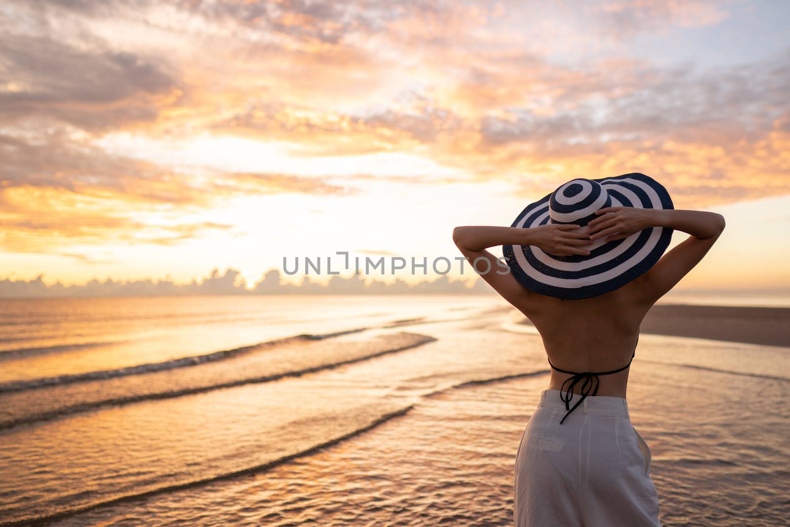 Woman in top bikini and white long pant wearing hat on the beach with a beautiful sunrise or sunset in background.