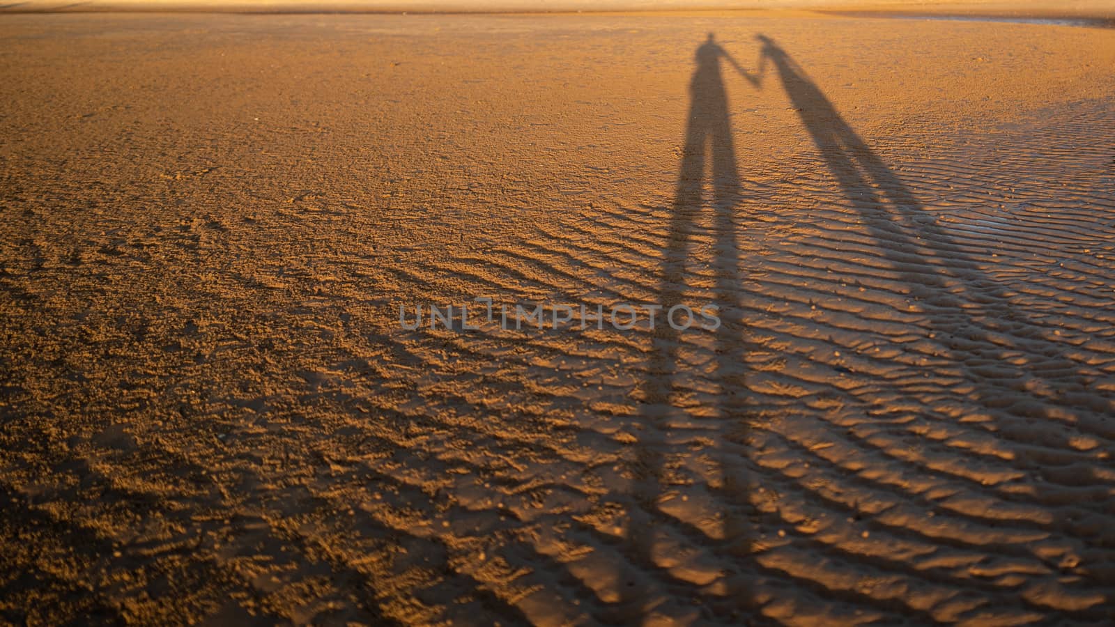 The shadow of a very long couple holding hand on a sand beach surface.