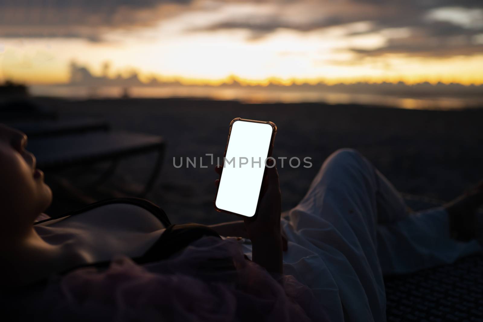 Woman hand holding mobile phone with blank white screen mockup on the beach in the morning.