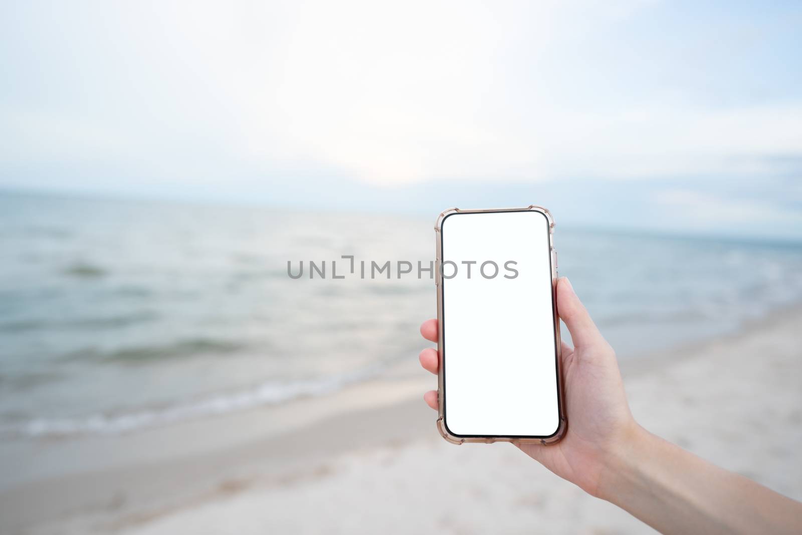 Woman hand holding mobile phone with blank white screen mockup on the beach.