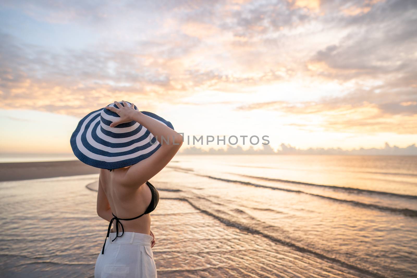 Woman in top bikini and white long pant wearing hat on the beach with a beautiful sunrise or sunset in background.