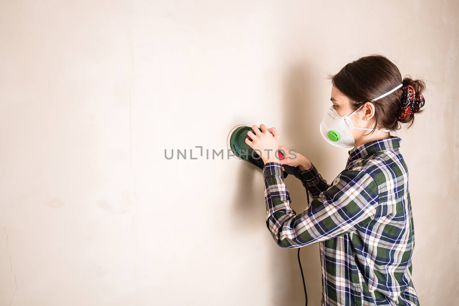 Woman in protective mask working with electric sander to smooth wall by Frostroomhead