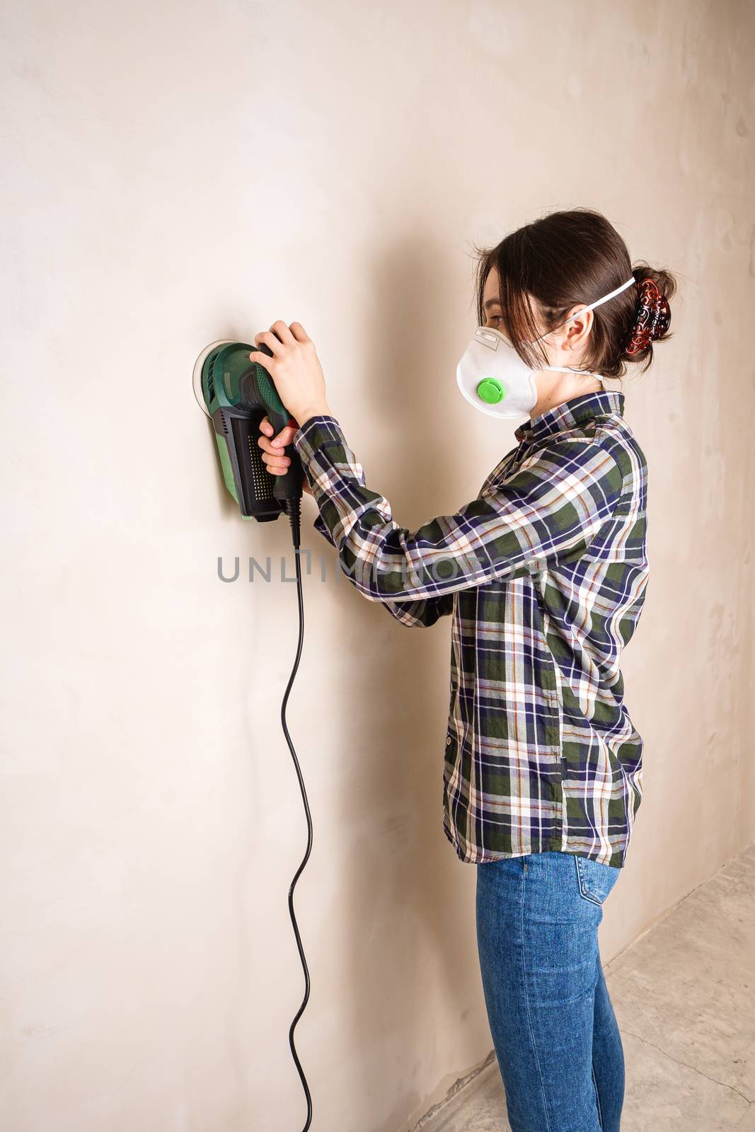 Woman in protective mask working with electric sander to smooth plaster wall surface, room renovation concept