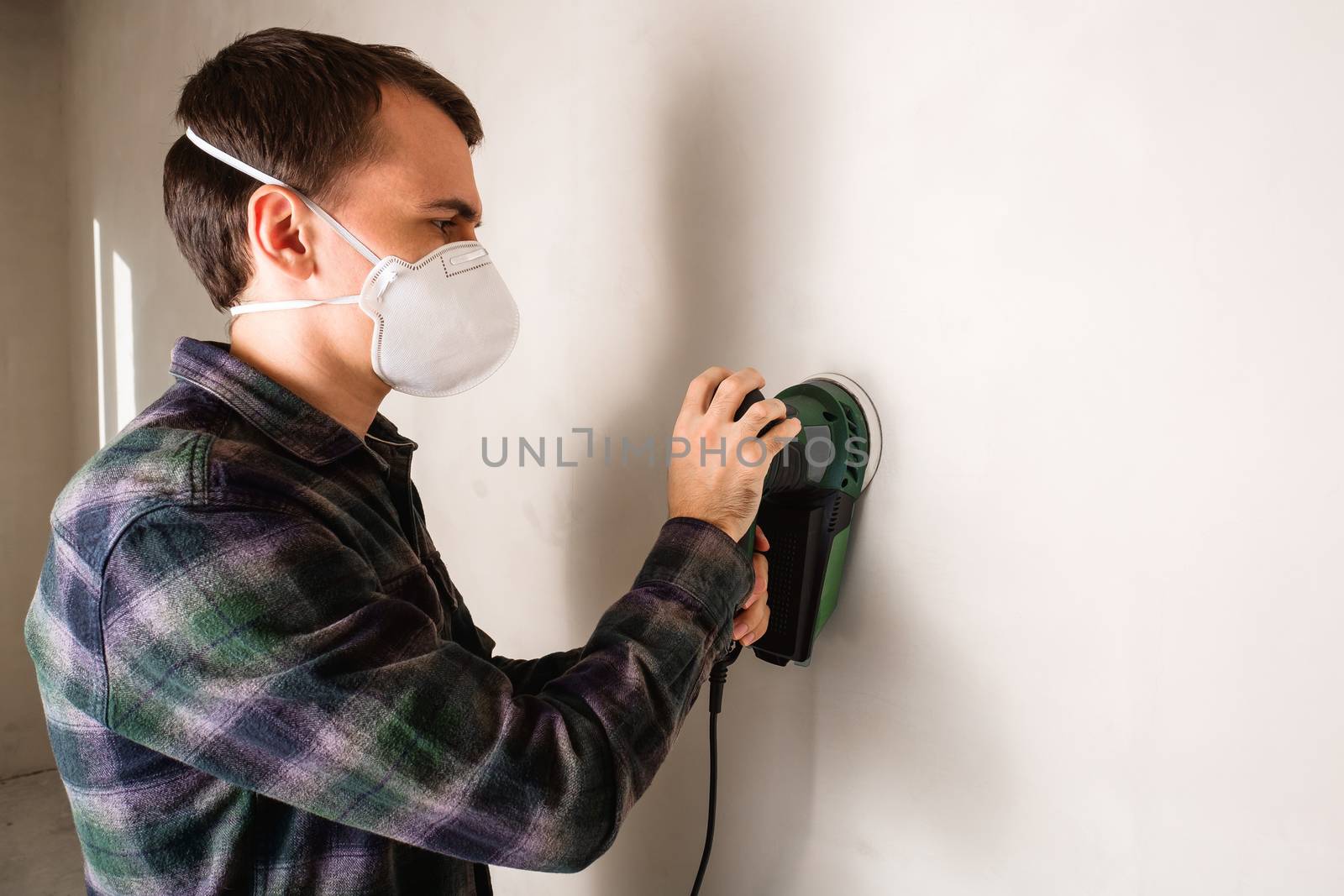 Man in protective mask working with electric sander to smooth plaster wall surface, room renovation concept