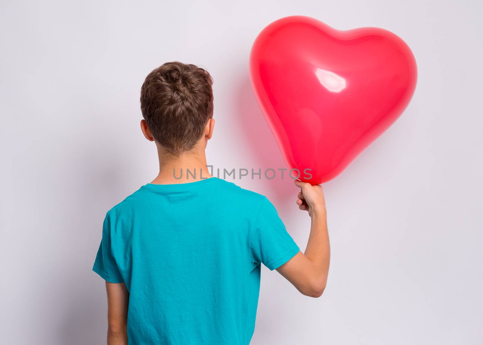 Back view. Portrait of teen boy holds red heart shaped balloon, on grey background. Child in blue t-shirt holding symbol of love, family, hope - rear view. Valentines Day concept.