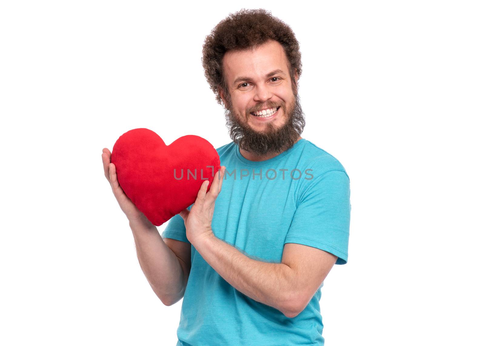 Happy Valentines Day. Crazy bearded Man with funny Curly Hair in blue T-shirt. Happy and silly guy in Love, isolated on white background. Portrait of Cheerful man with Red plush Heart, look at camera