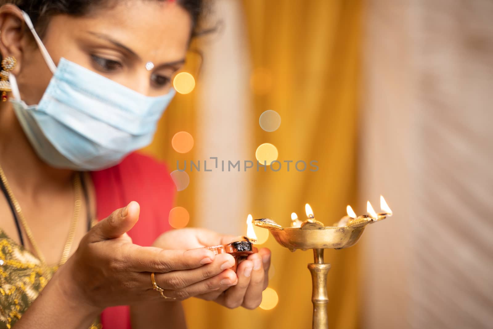 Selective focus on hands, Indian woman in medical mask lighting lantern or Diya Lamp during festival at home - concept of traditional festival celebrations during Coronavirus or Covid-19 Pandemic by lakshmiprasad.maski@gmai.com