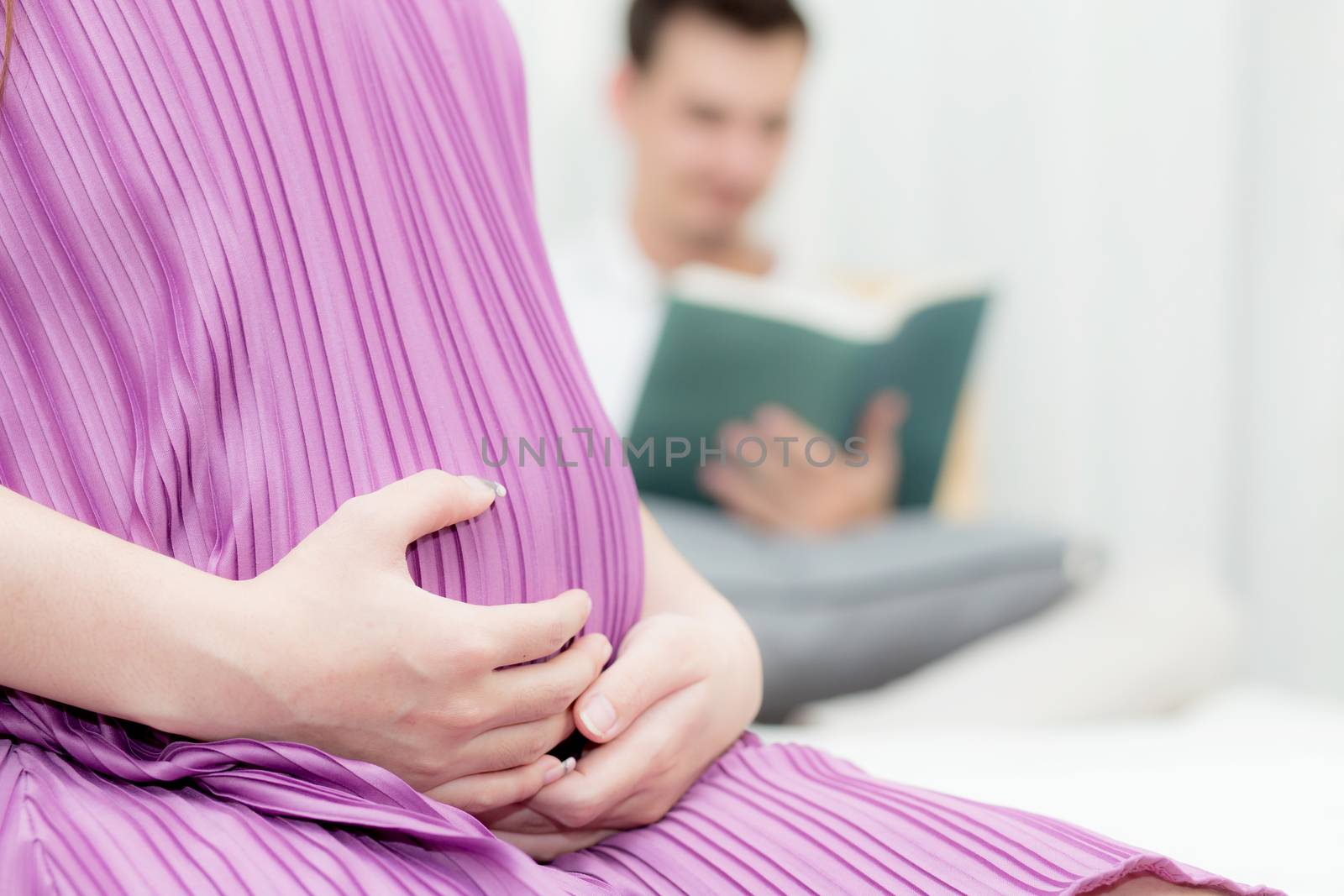 Asian pregnant people and expectation concept - happy pregnant woman sitting on bed and touching her belly at home with father reading book background.