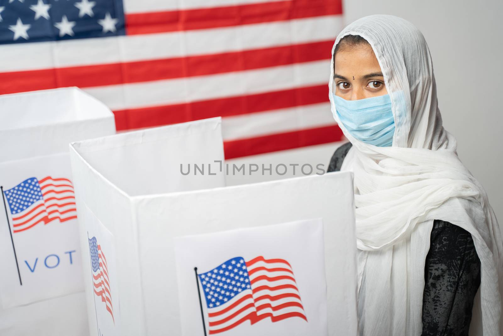 Girl with Hijab or head covering and mask worn at polling booth looking at camera with US flag as background - Concept of US election. by lakshmiprasad.maski@gmai.com