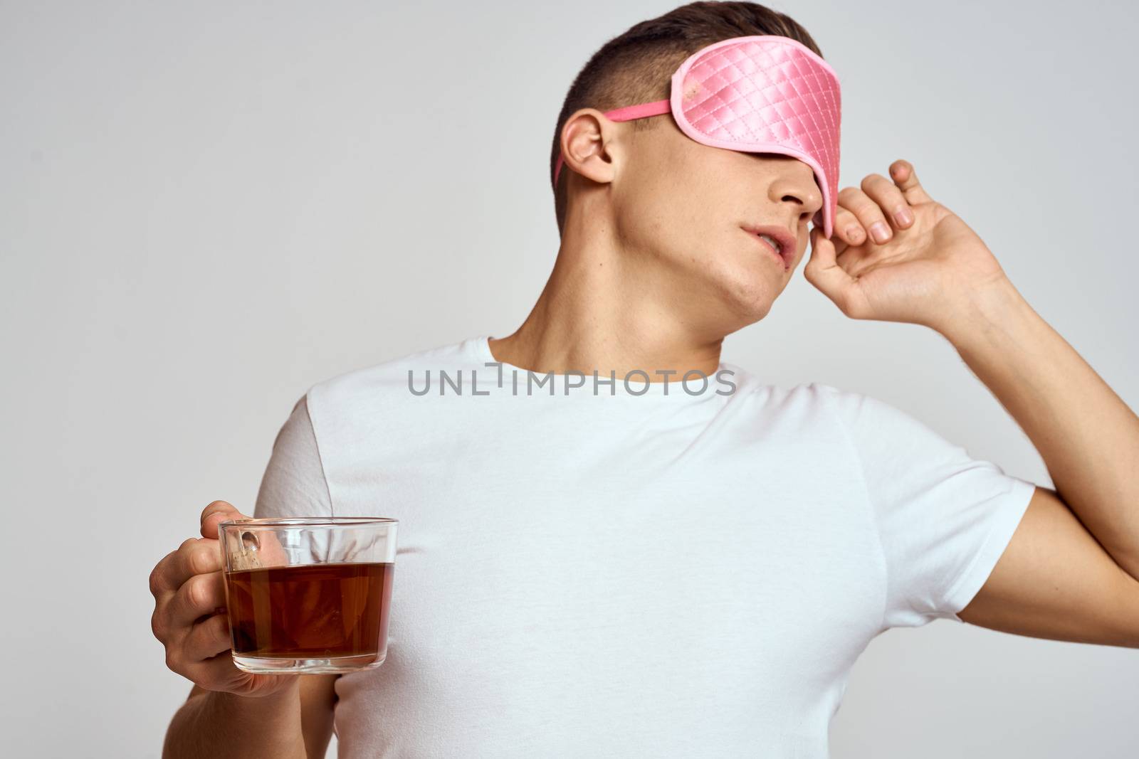 handsome man with pink sleep mask and with a cup of tea in hand pulls himself up on a light background cropped view model. High quality photo