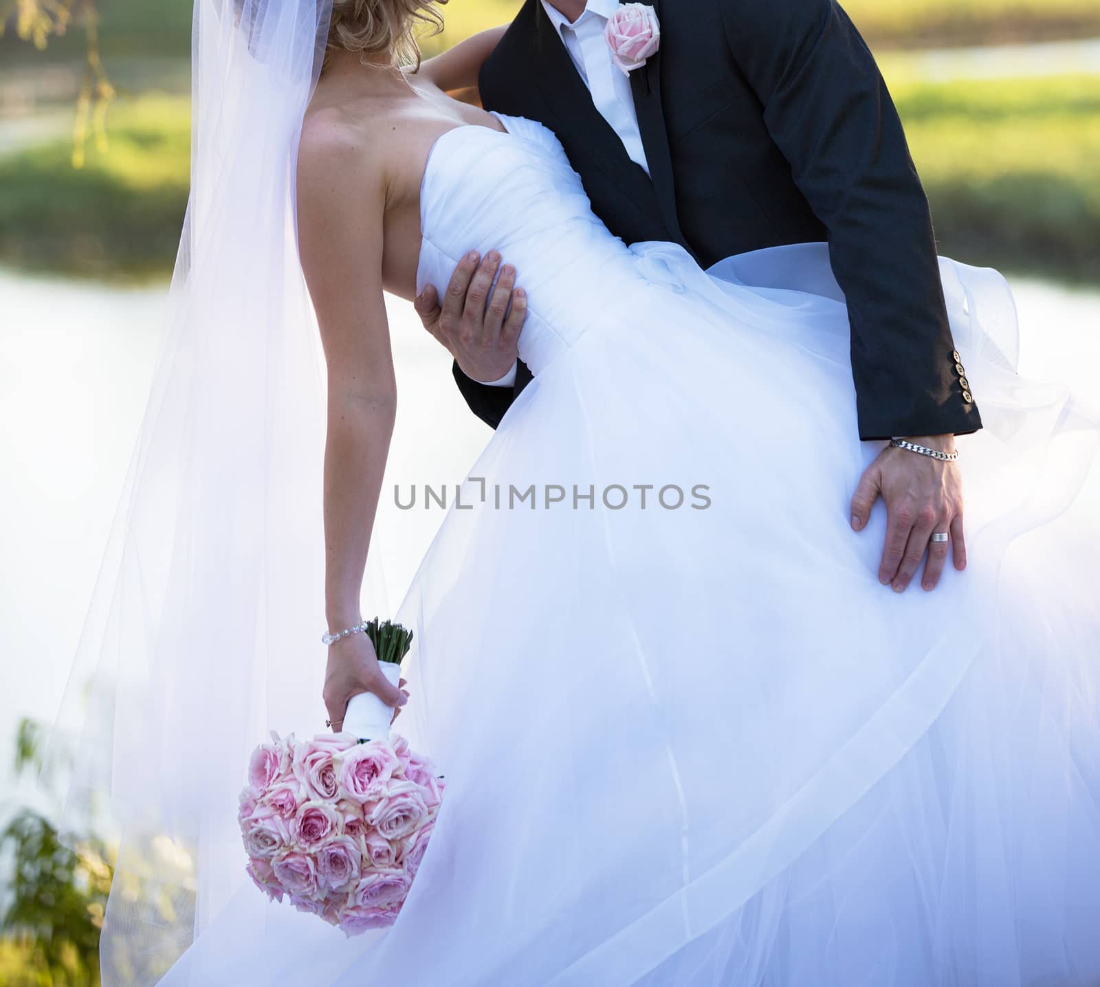 The groom leans his bride backwards for a kiss beside a lake in soft afternoon light