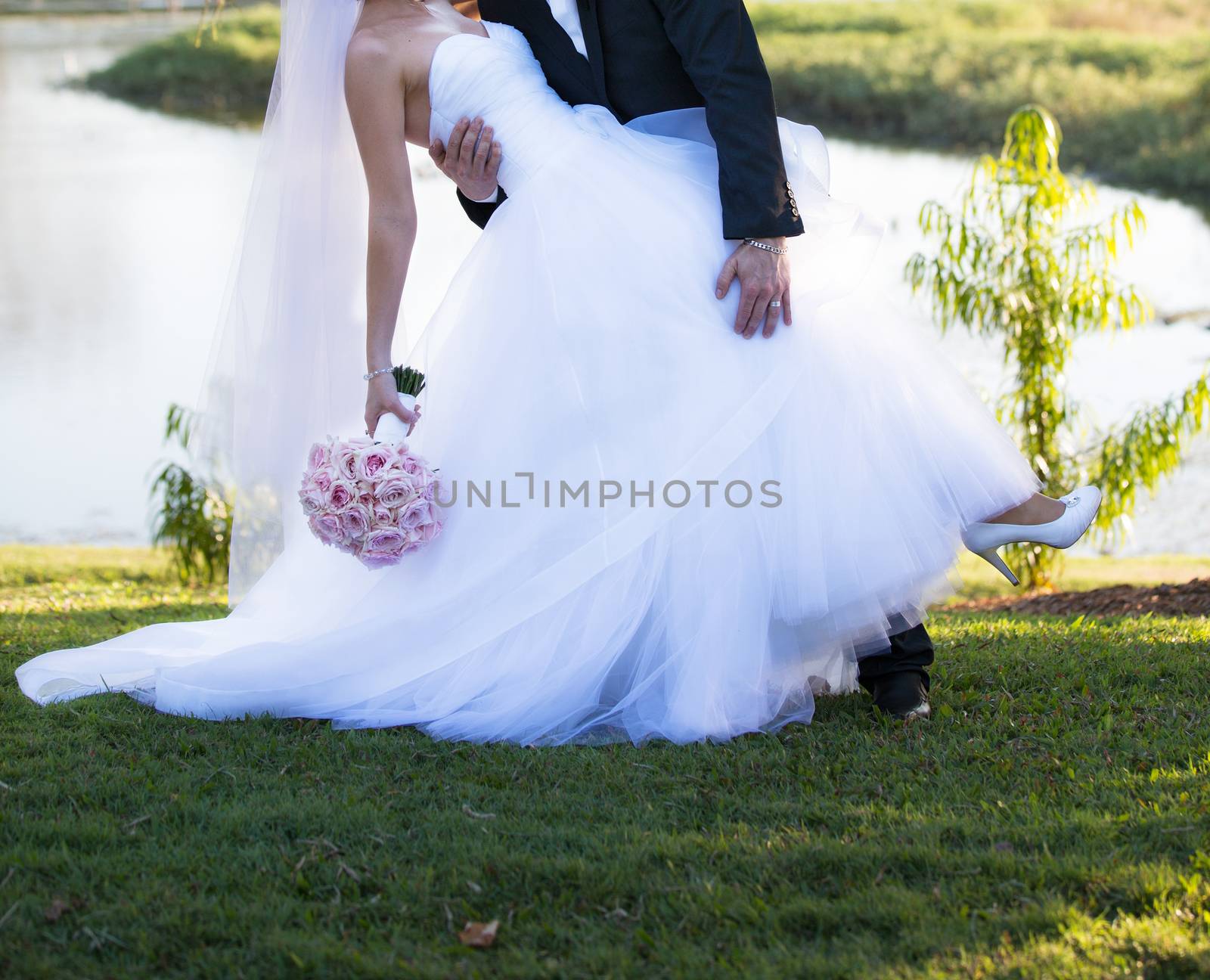 The groom leans his bride backwards for a kiss beside a lake in soft late afternoon light