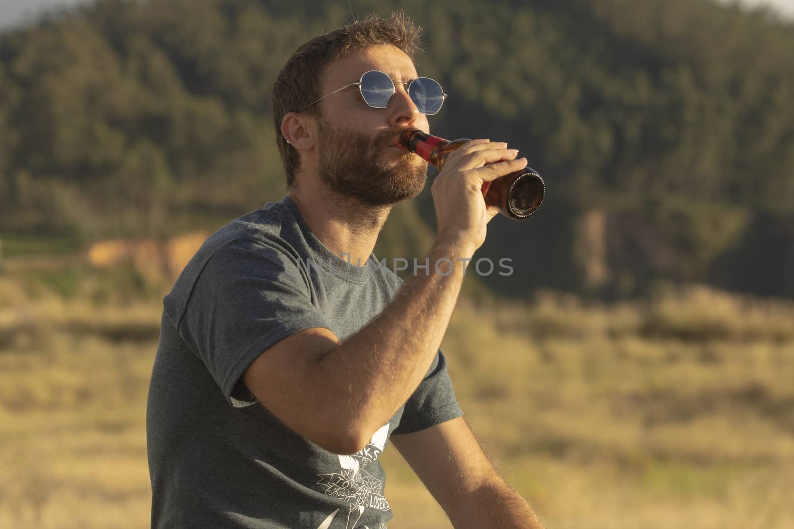 A young man, with glasses and a beard, drinks beer from a bottle, while enjoying the landscape and the sunset, in Galicia, northern Spain.