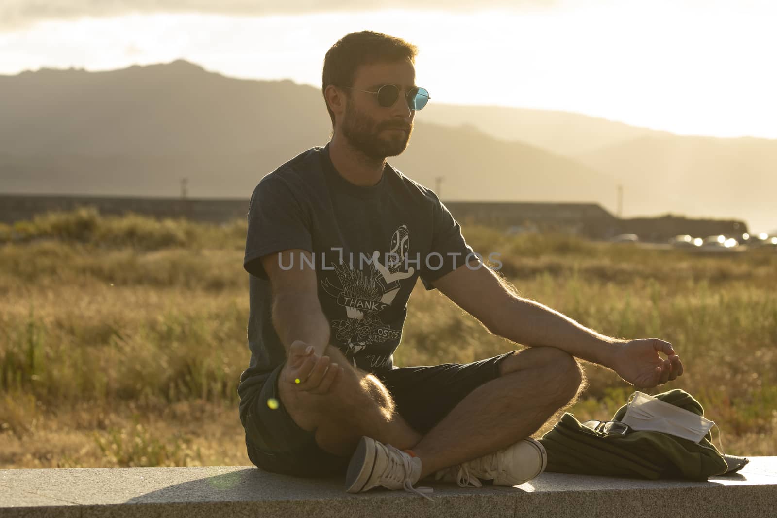 A young man, wearing glasses, meditates in a yoga posture, while enjoying the afternoon sun on a promenade in northern Spain.