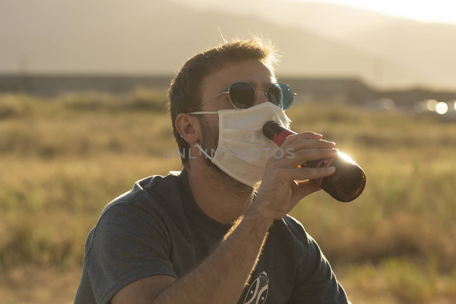 A young man, with glasses, tries to drink a beer wearing a protective face mask.