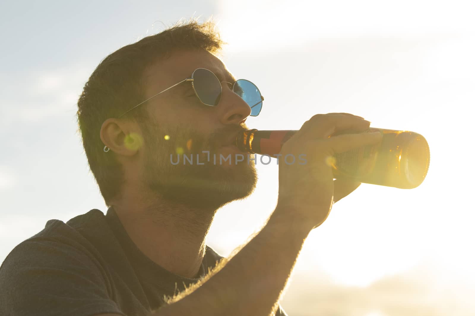 A young man, with glasses and a beard, drinks beer from a bottle, while enjoying the landscape and the sunset, in Galicia, northern Spain.