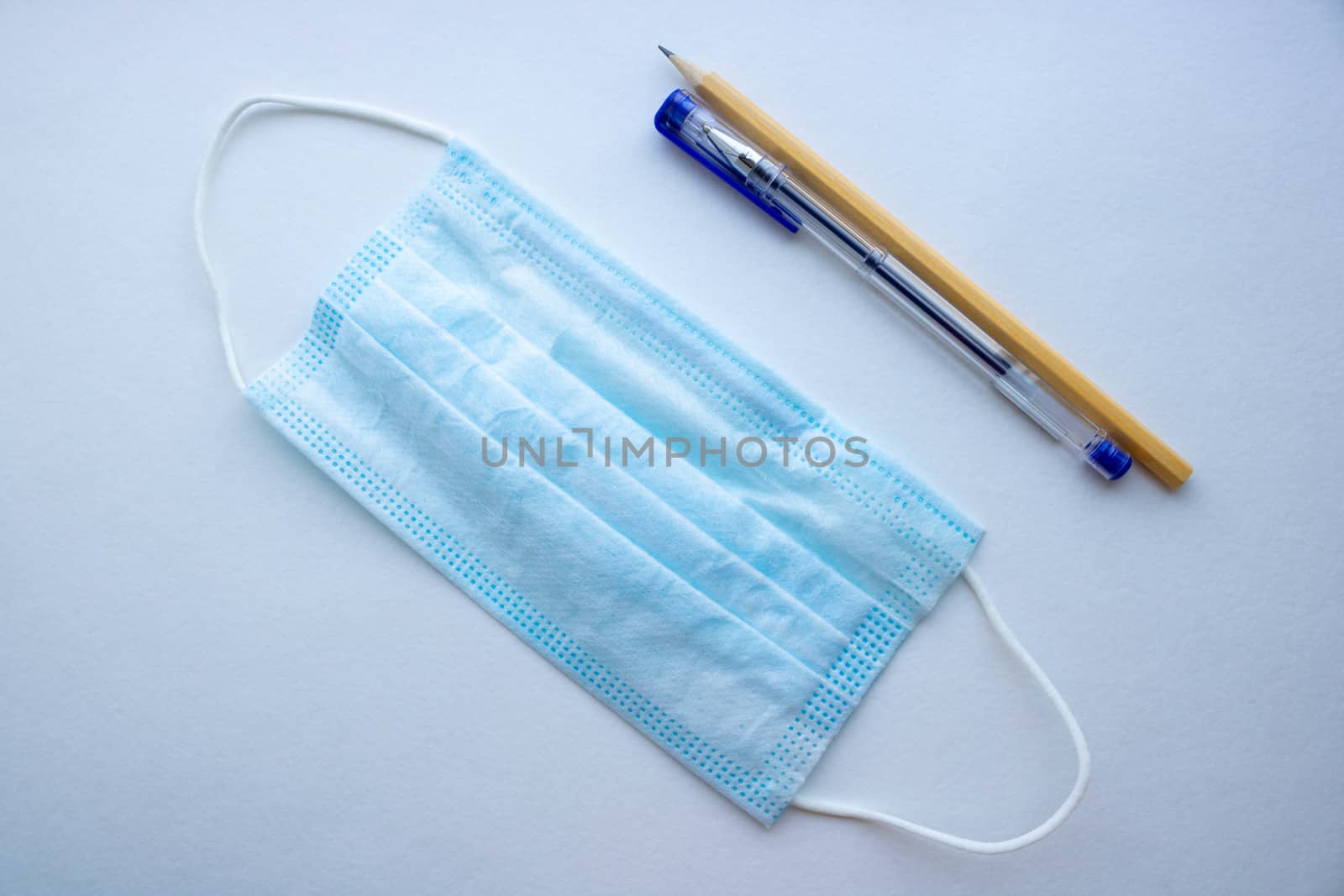 A surgical or medical mask with rubber ear straps lies next to a pen and pencil on a white background. A typical 3-layer doctor's mask to cover your mouth and nose.The concept of protection.