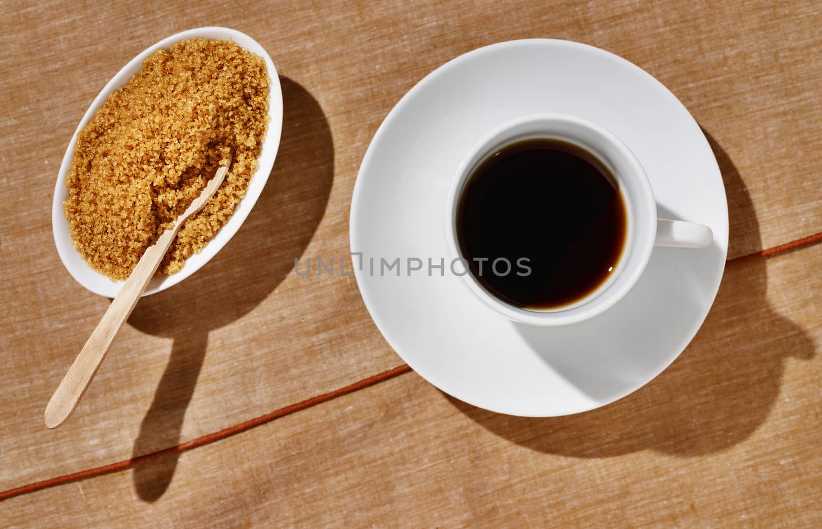 A white cup of coffee with white sugar bowl with brown sugar and spoon , brown table cloth