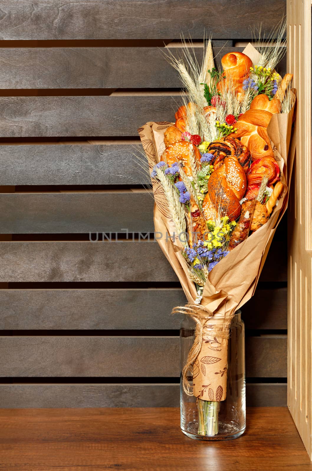Fresh baked goods and bread, wheat ears and wildflowers are beautifully wrapped in a bouquet of craft paper, standing in a glass vase on a wooden striped brown background, copy space.