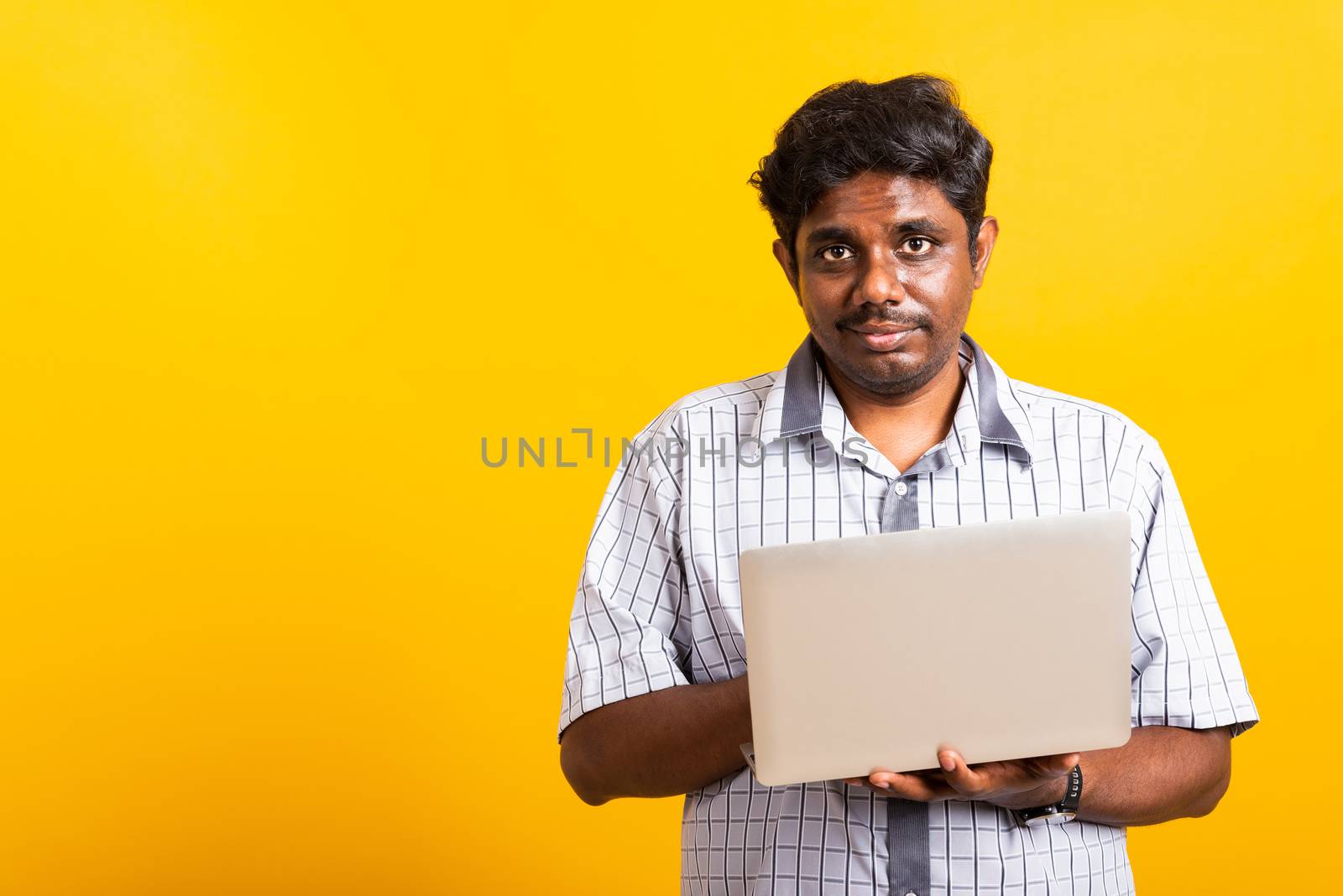 Asian happy portrait young black man smiling standing wear shirt holding and typing laptop computer he looking to camera isolated, studio shot yellow background with copy space