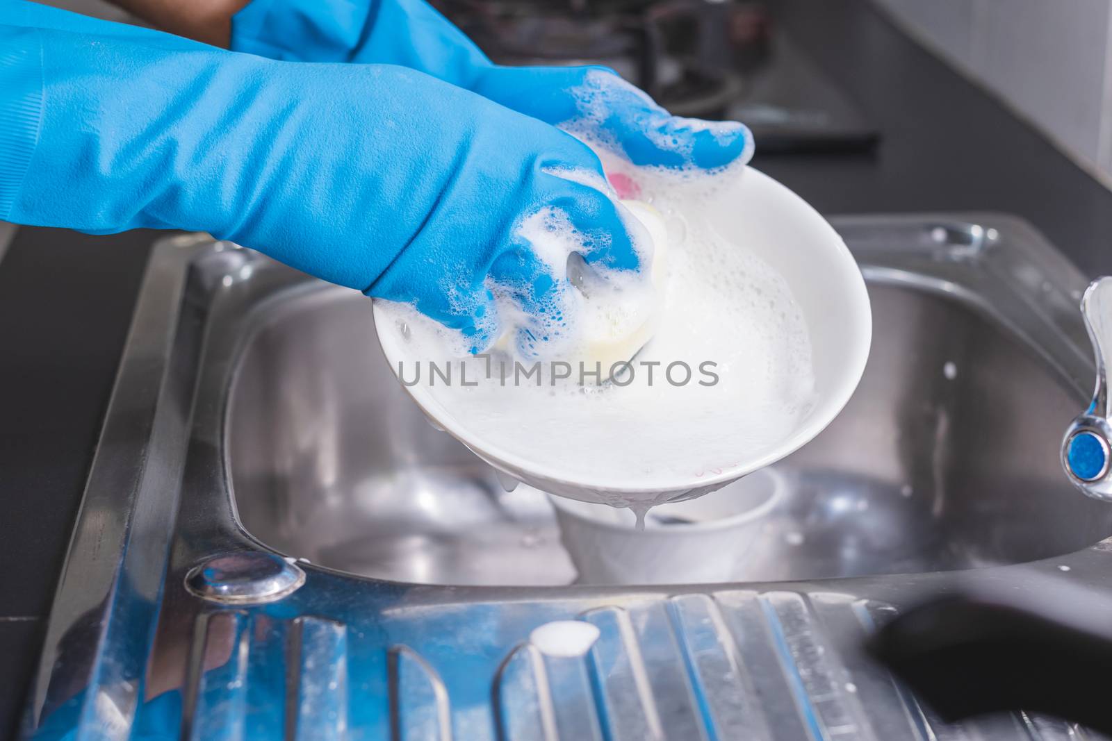 Close up of a man wearing a blue rubber glove was washing the cup with a dish washing liquid in the sink in the kitchen.