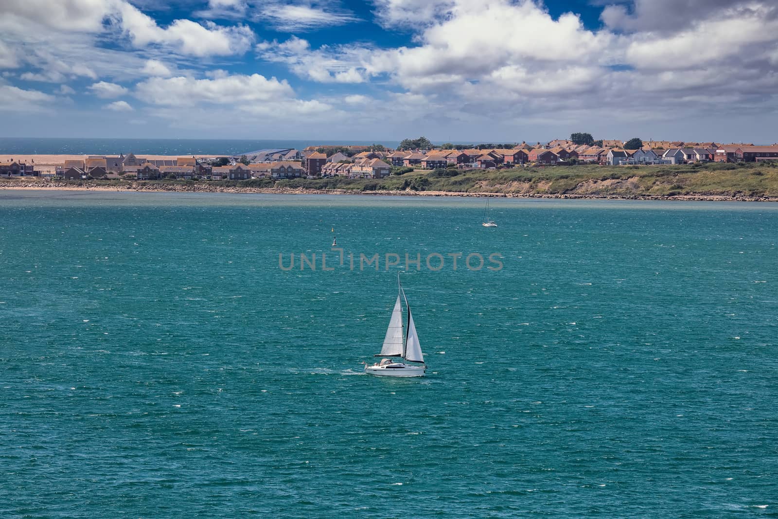 High angle shot of a white sailboat sailing in Weymouth Bay, UK. Coast line with some houses and gorgeous cloudy blue sky in the background. Sport and recreation concept.