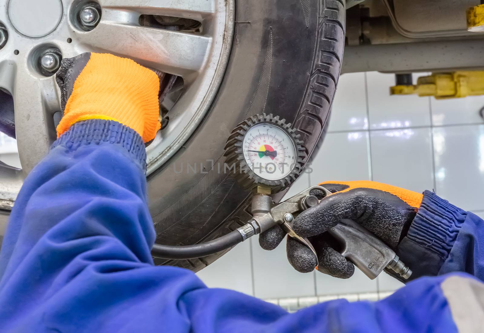 Closeup of mechanic wearing orange gloves and blue outfit at repair service station checking tyre pressure with gauge.