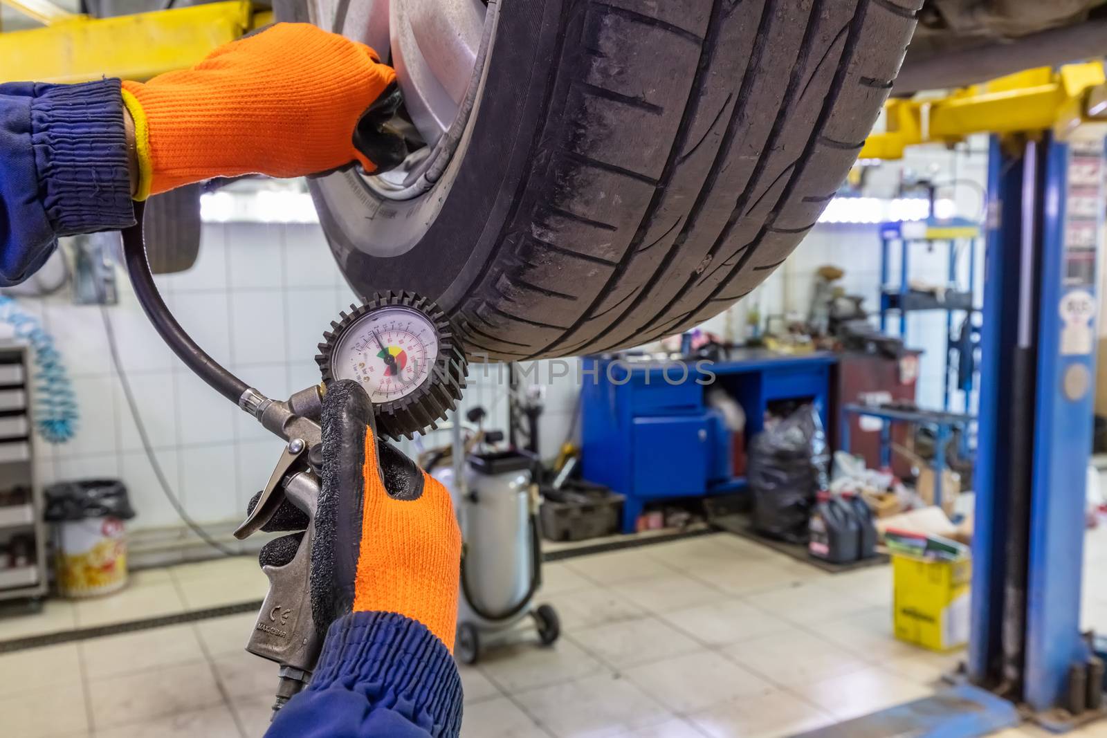 Closeup of mechanic wearing orange gloves and blue outfit at repair service station checking tyre pressure with gauge.