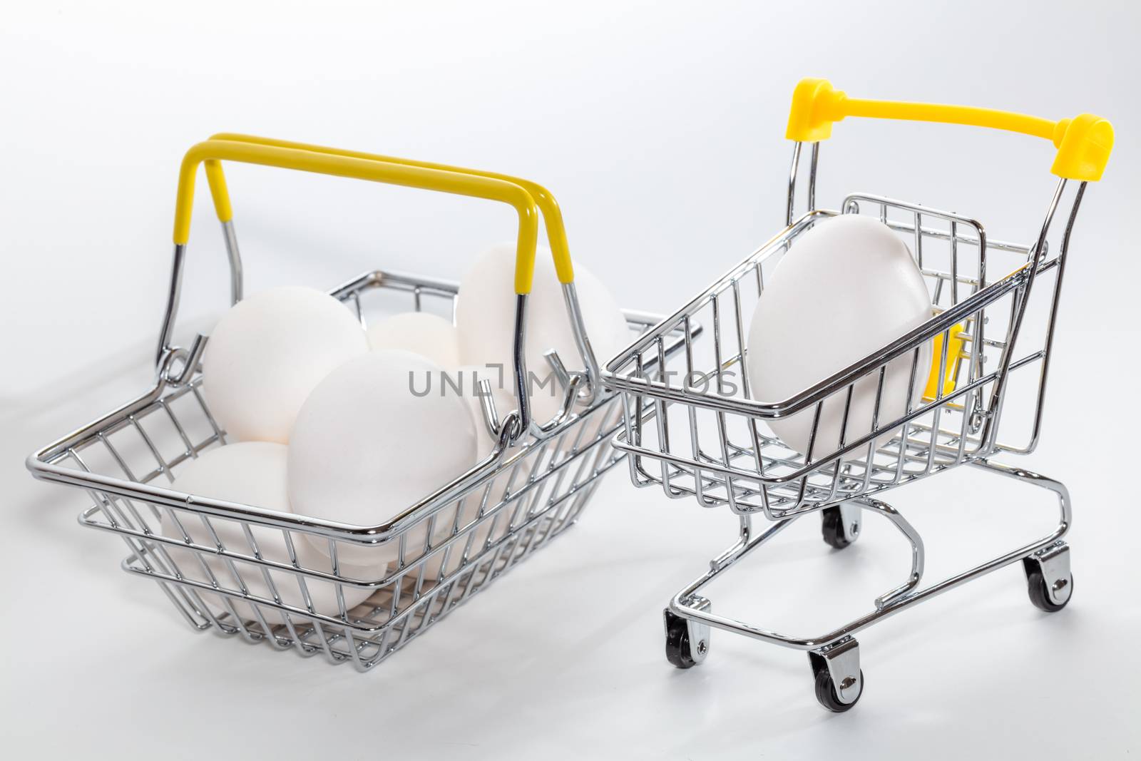Fresh eggs in a shopping cart and a basket next to it. Eggs in a basked slightly out of focus. High angle view. Shopping, purchasing, and food delivery concept. White background. Close up shot. Isolated.