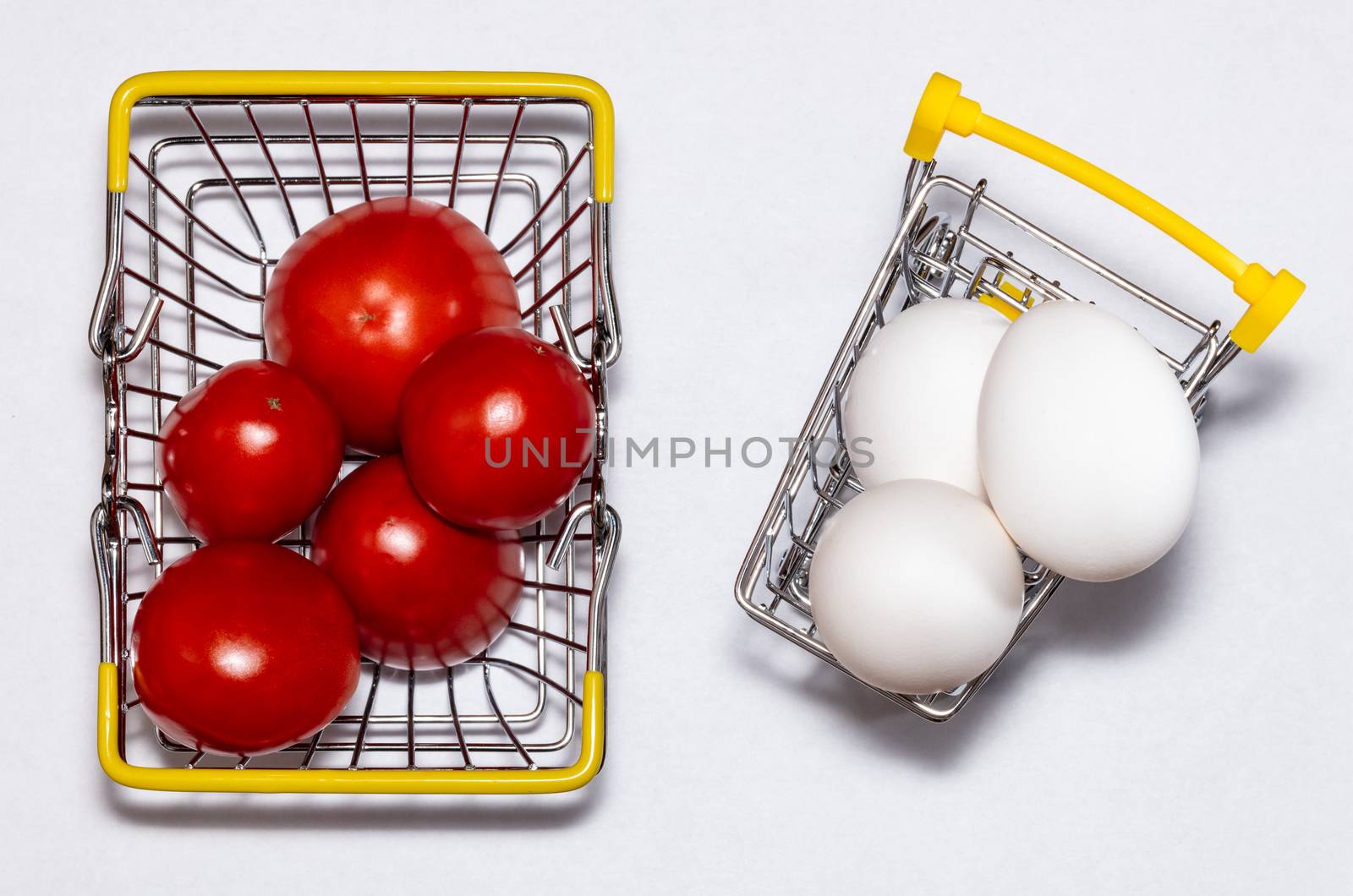 Fresh eggs and tomatoes in a shopping cart and a basket next to it. All mixed up. Top view. Shopping, purchasing, and food delivery concept. White background. Close up shot. Isolated.