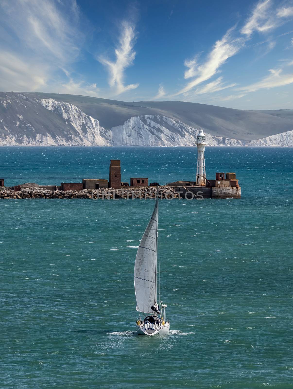 High angle shot of a white sailboat sailing in Weymouth Bay, UK. Coast line with white cliffs and gorgeous cloudy blue sky in the background. Lighthouse at the harbour entrance. Sport and recreation concept. Vertical, portrai format.
