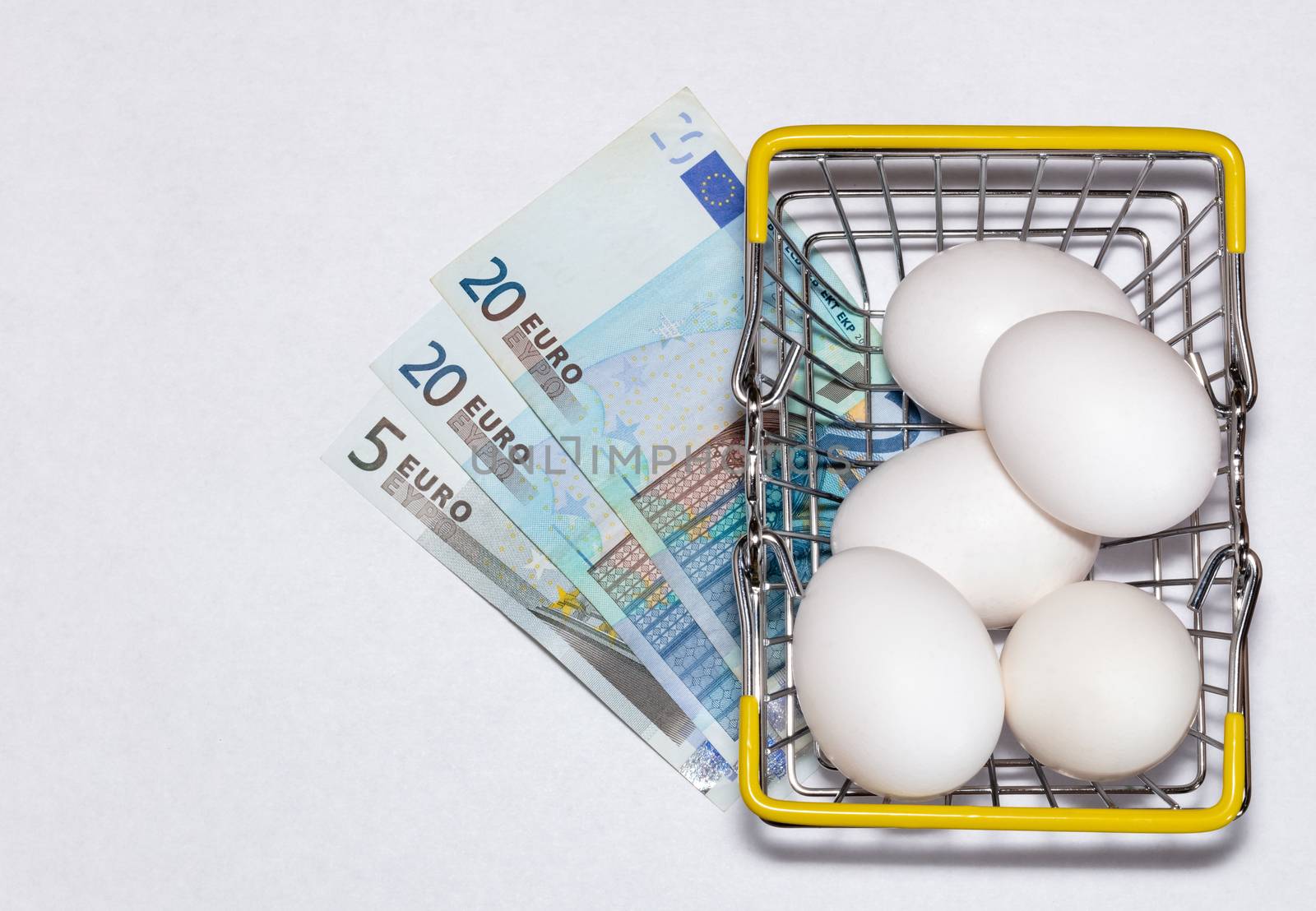 Fresh eggs in a shopping cart with various euro bills underneath it. Shopping, purchasing, and food delivery concept. White background. Close up shot. Isolated.