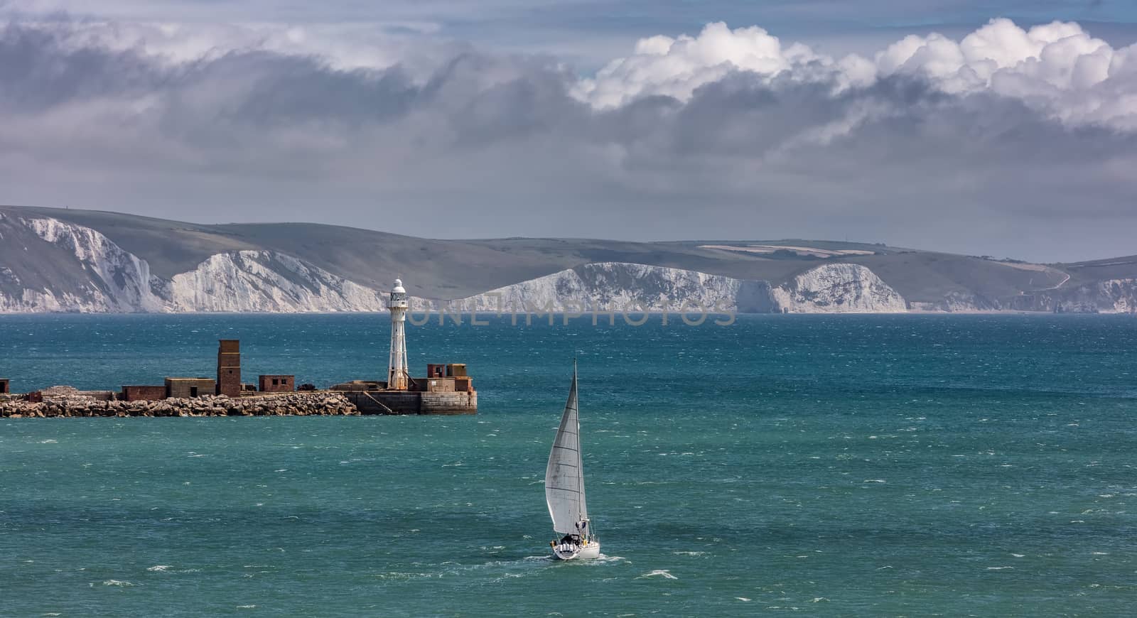 High angle shot of a white sailboat sailing in Weymouth Bay, UK. Coast line with white cliffs and cloudy sky in the background. Lighthouse at the harbour entrance. Sport and recreation concept. Panorama size.