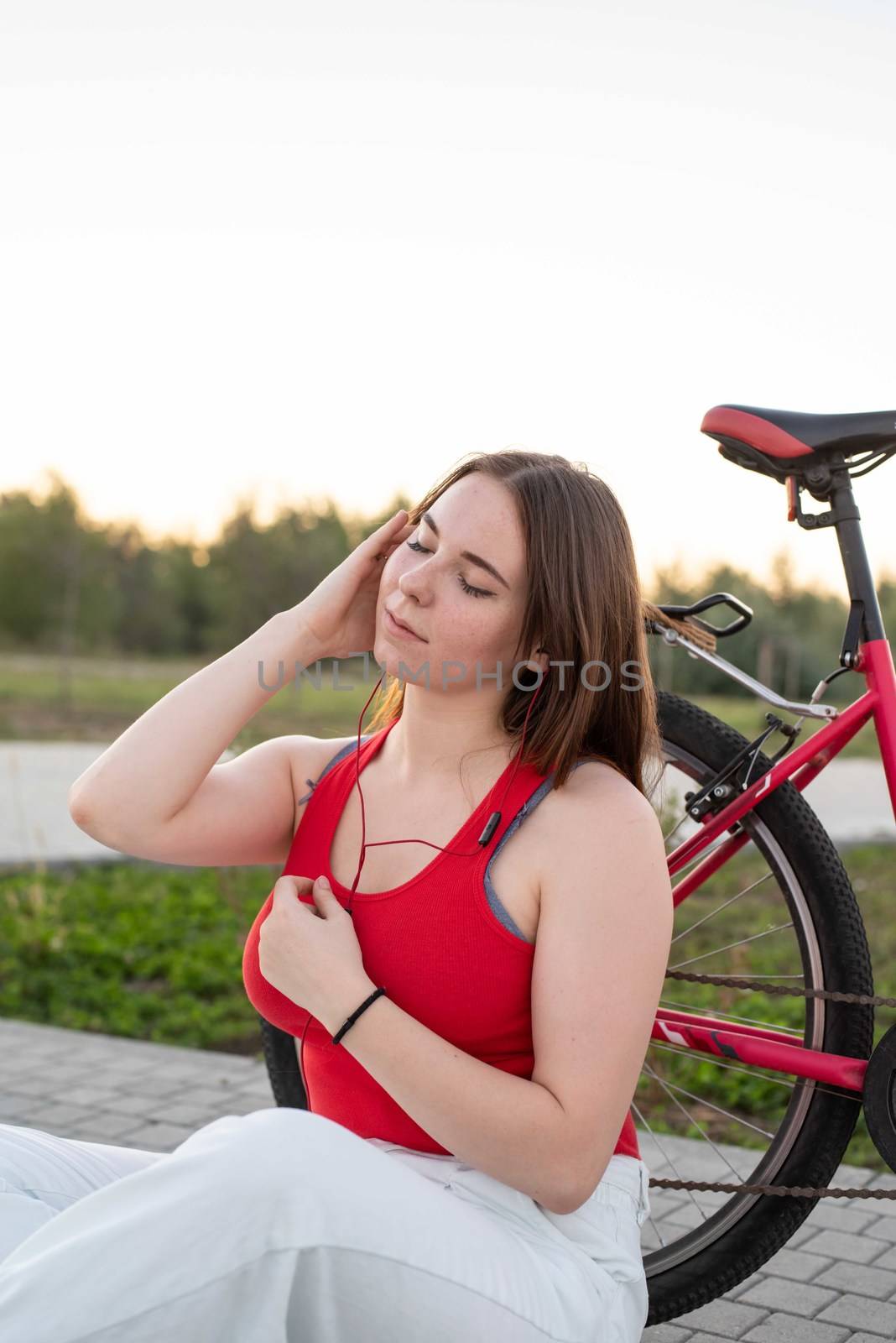 Teenage girl sitting next to her bike listening to the music in the park at sunset by Desperada