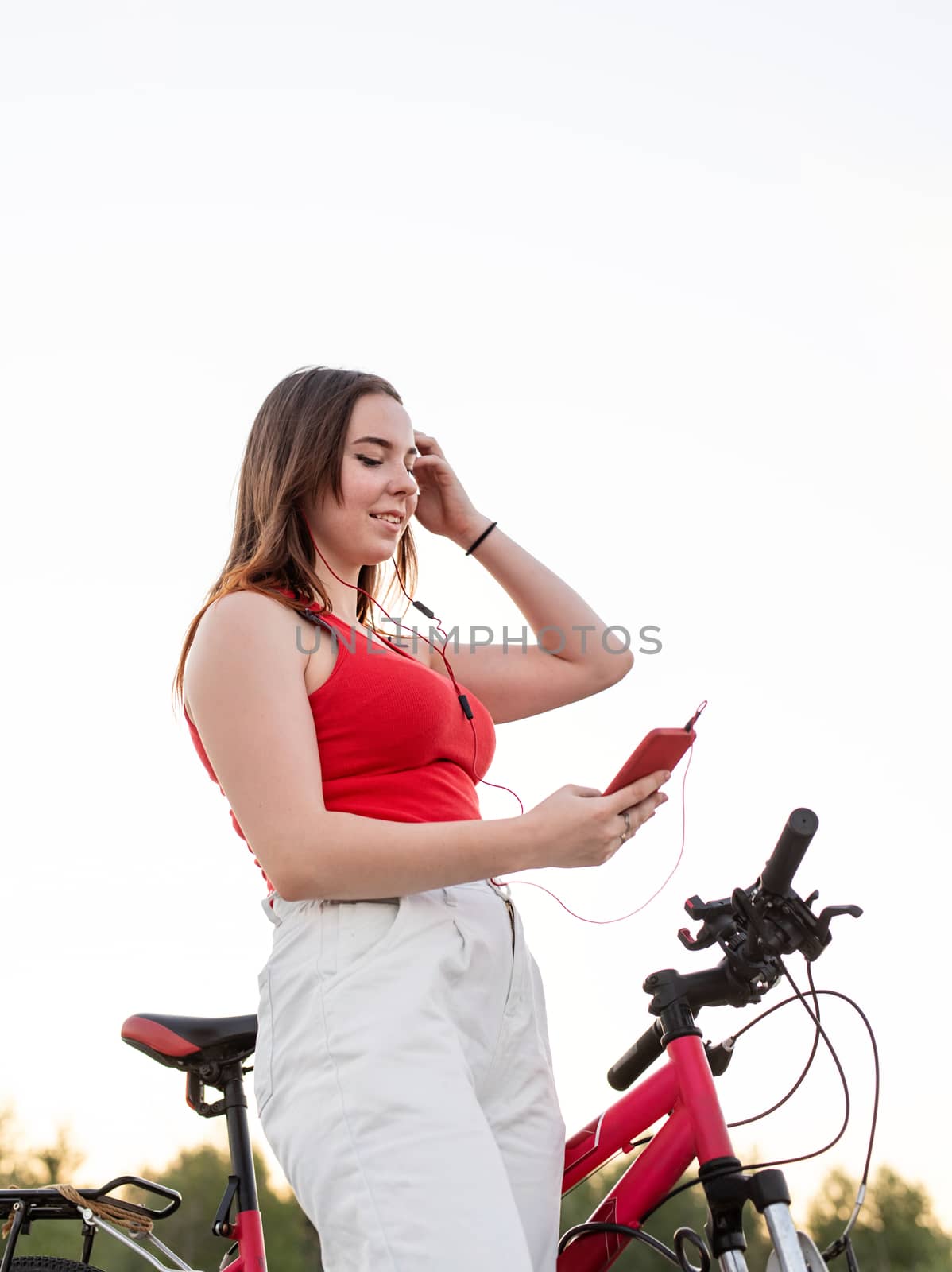 Teenage girl standing next to her bike listening to the music in headphones in the park at sunset. Active lifestyle