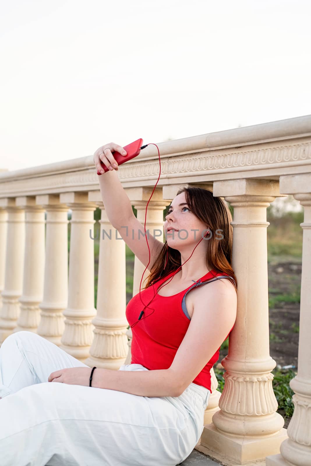 Teenage girl sitting next to column making selfie and listening to the music in the park at sunset by Desperada
