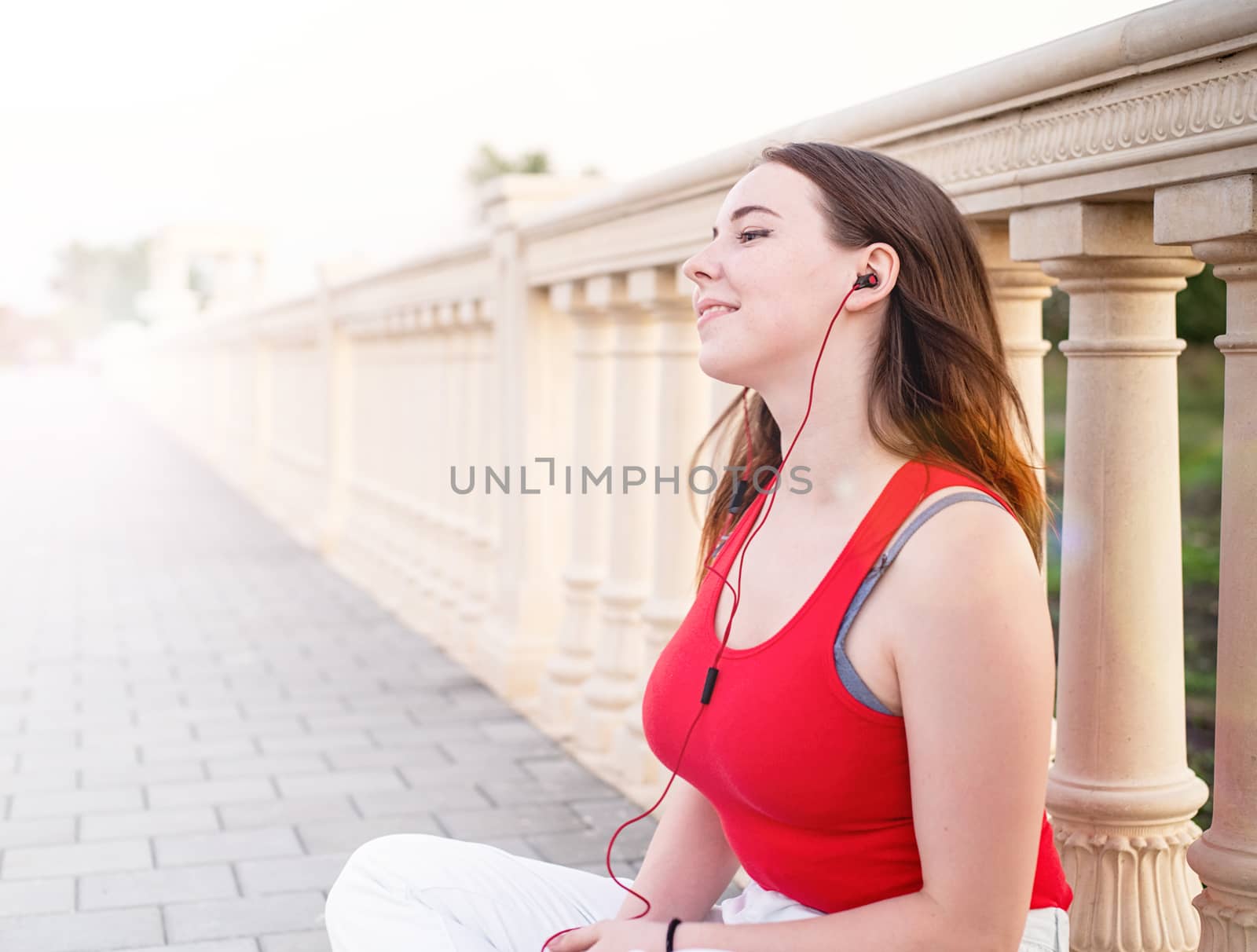 Teenage girl sitting next to column fencing listening to the music in the park at sunset by Desperada