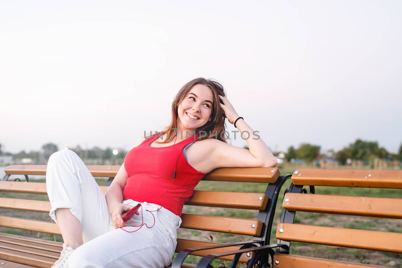 Happy young woman sitting on a bench in the park listening to the music in headphones