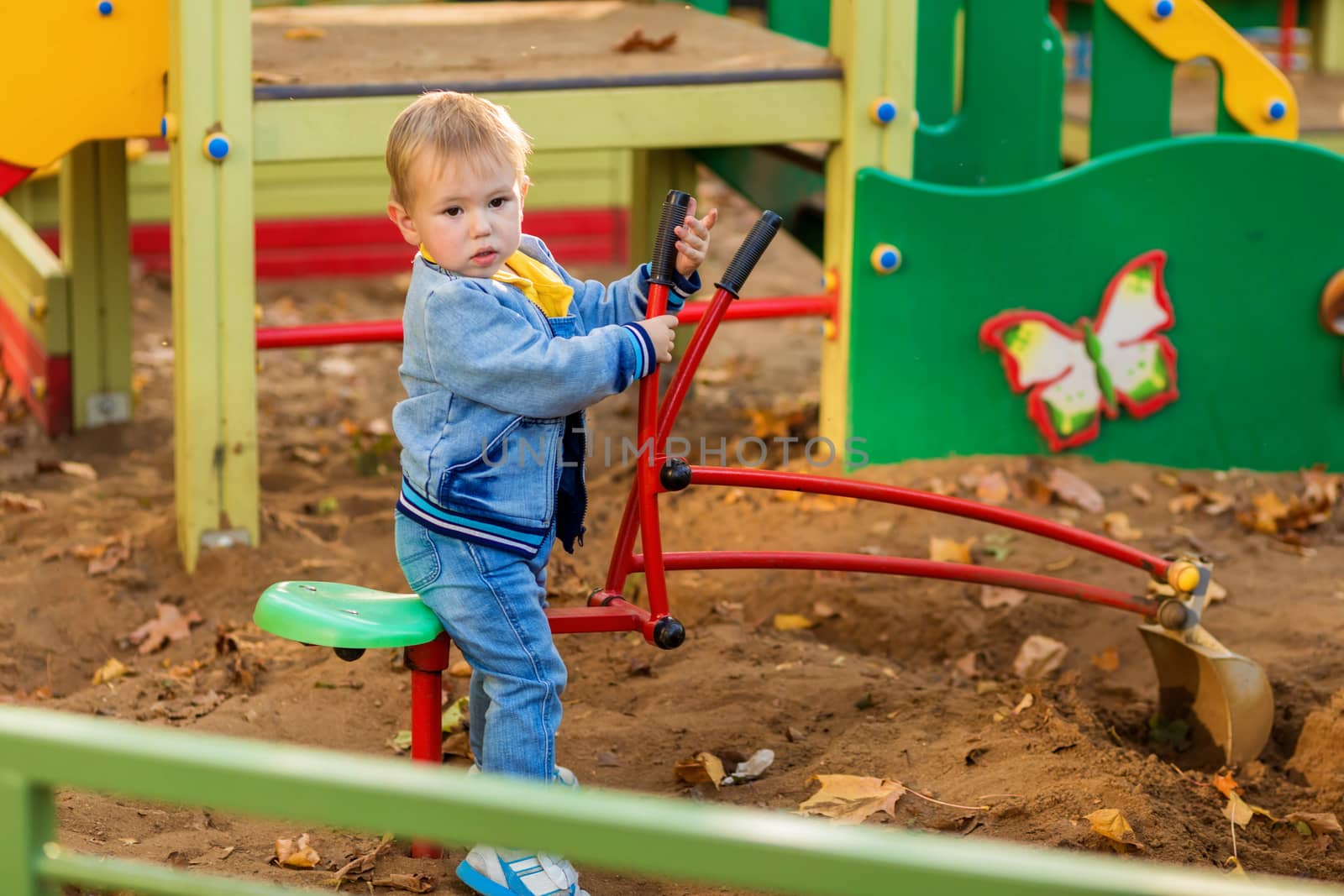 Little boy in denim clothes plays with a toy excavator on the playground in the autumn park.