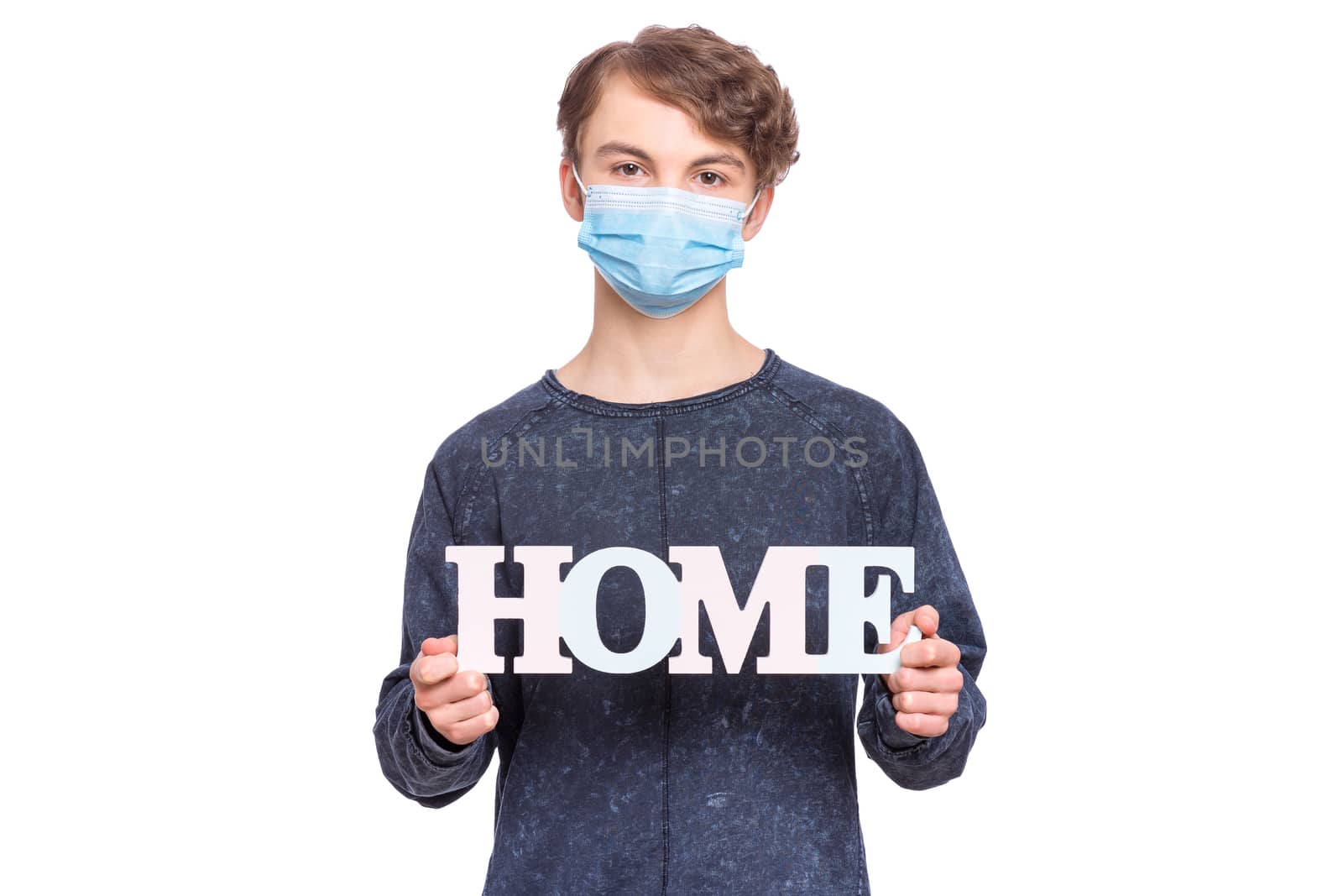 Concept of coronavirus quarantine. Child wearing medical protective face mask during flu virus, holds word Home - message for people to stay at home. COVID-19 - self isolation. Boy isolated on white.