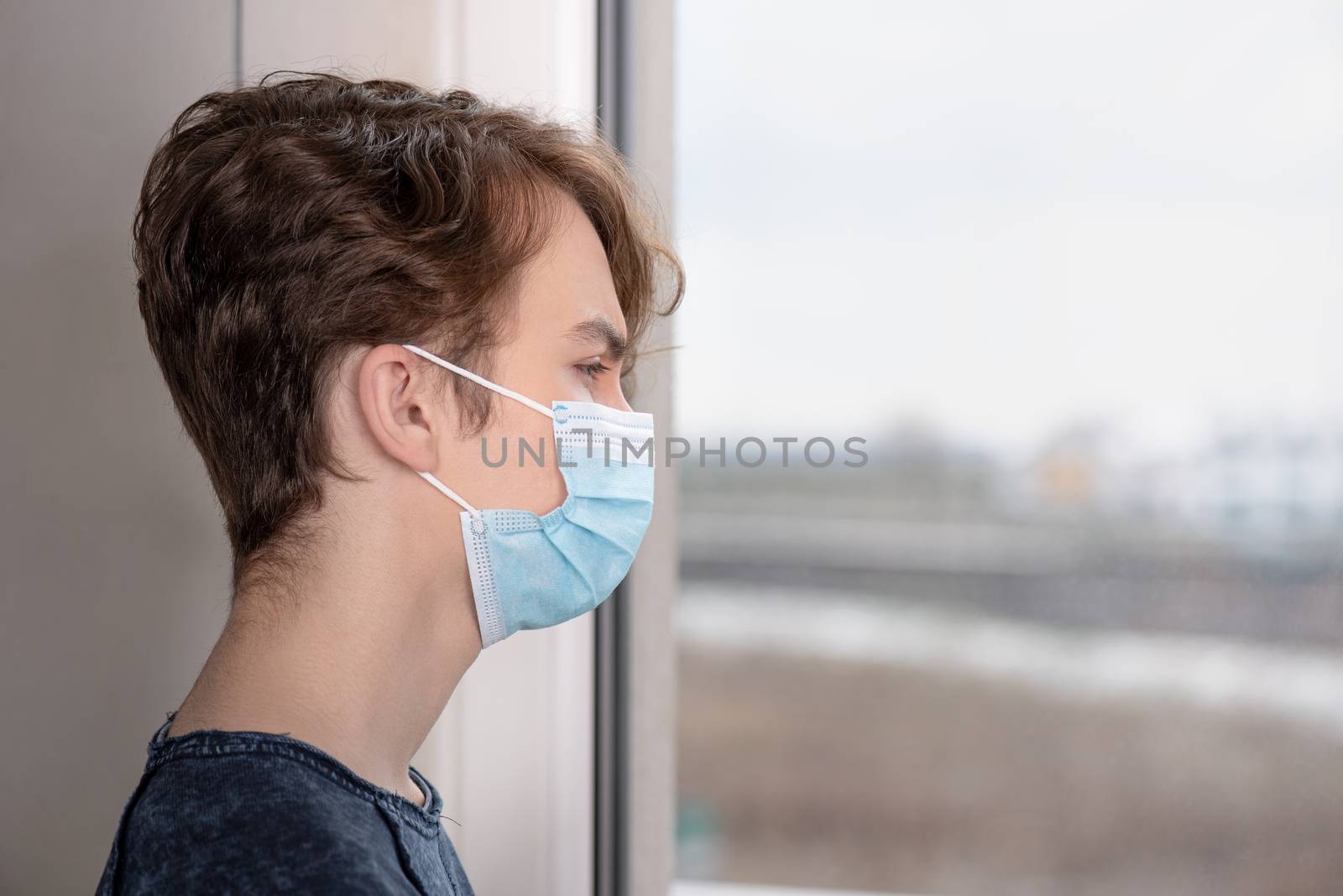 Concept of coronavirus quarantine. Child wearing medical protective face mask during flu virus, looking out of window. COVID-19 - self isolation. Teen boy forced to stay at home.