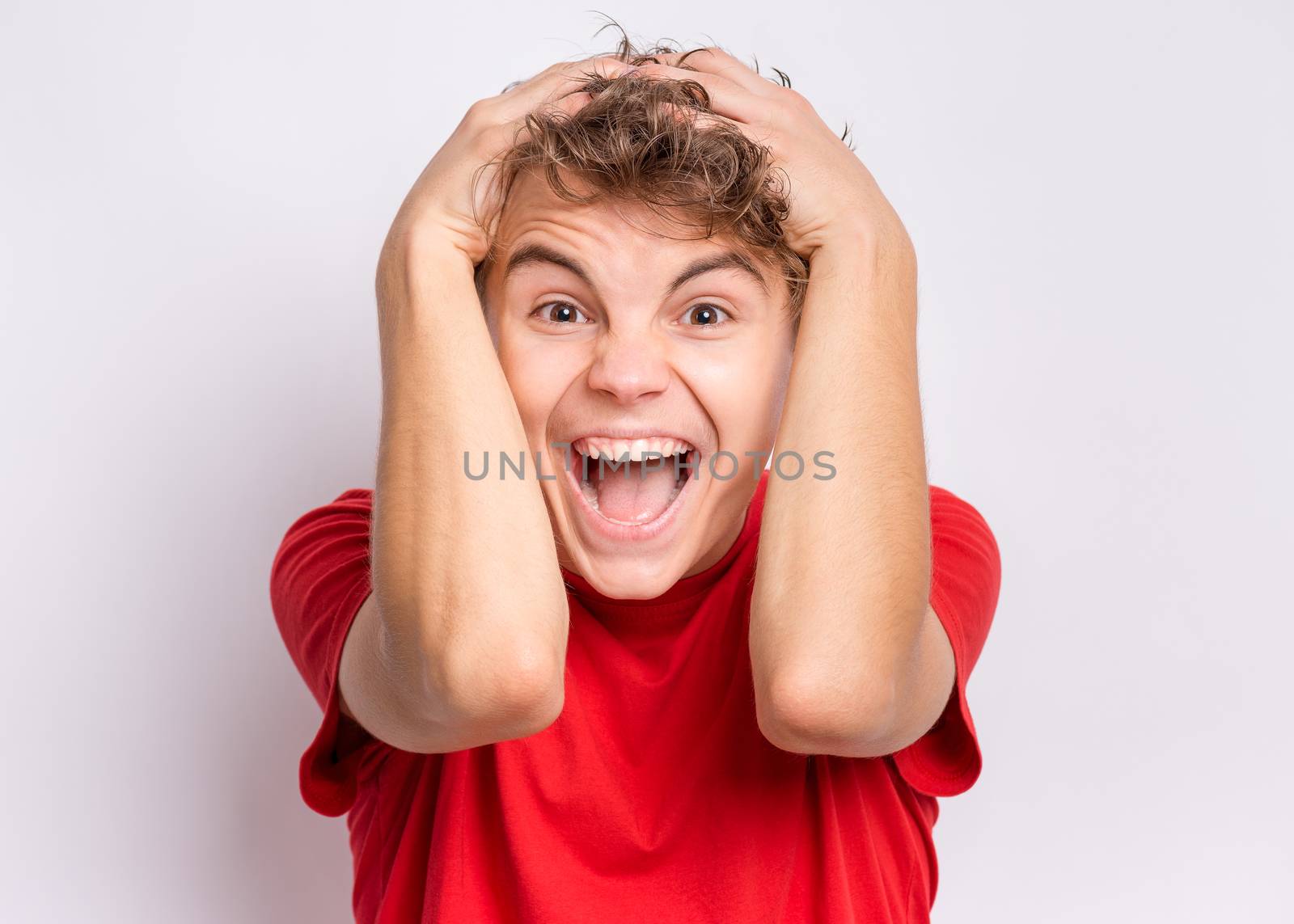 Portrait of surprised teen boy, on grey background. Funny child looking at camera in shock or amazement touching his head with hands. Handsome caucasian young teenager.