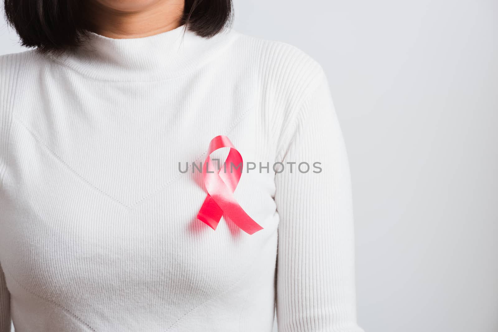 Breast cancer awareness healthcare and medicine concept. Close up Asian woman wear white shirt standing with pink breast cancer awareness ribbon pin on chest, studio shot isolated on white background