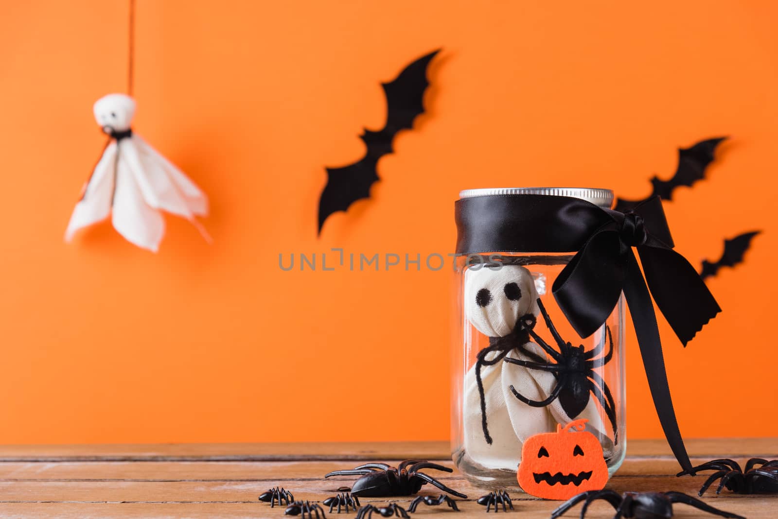 Funny Halloween day decoration party, Baby white ghost crafts scary face in jar glass on wood table, studio shot isolated on orange background have spider and bats, Happy holiday DIY concept