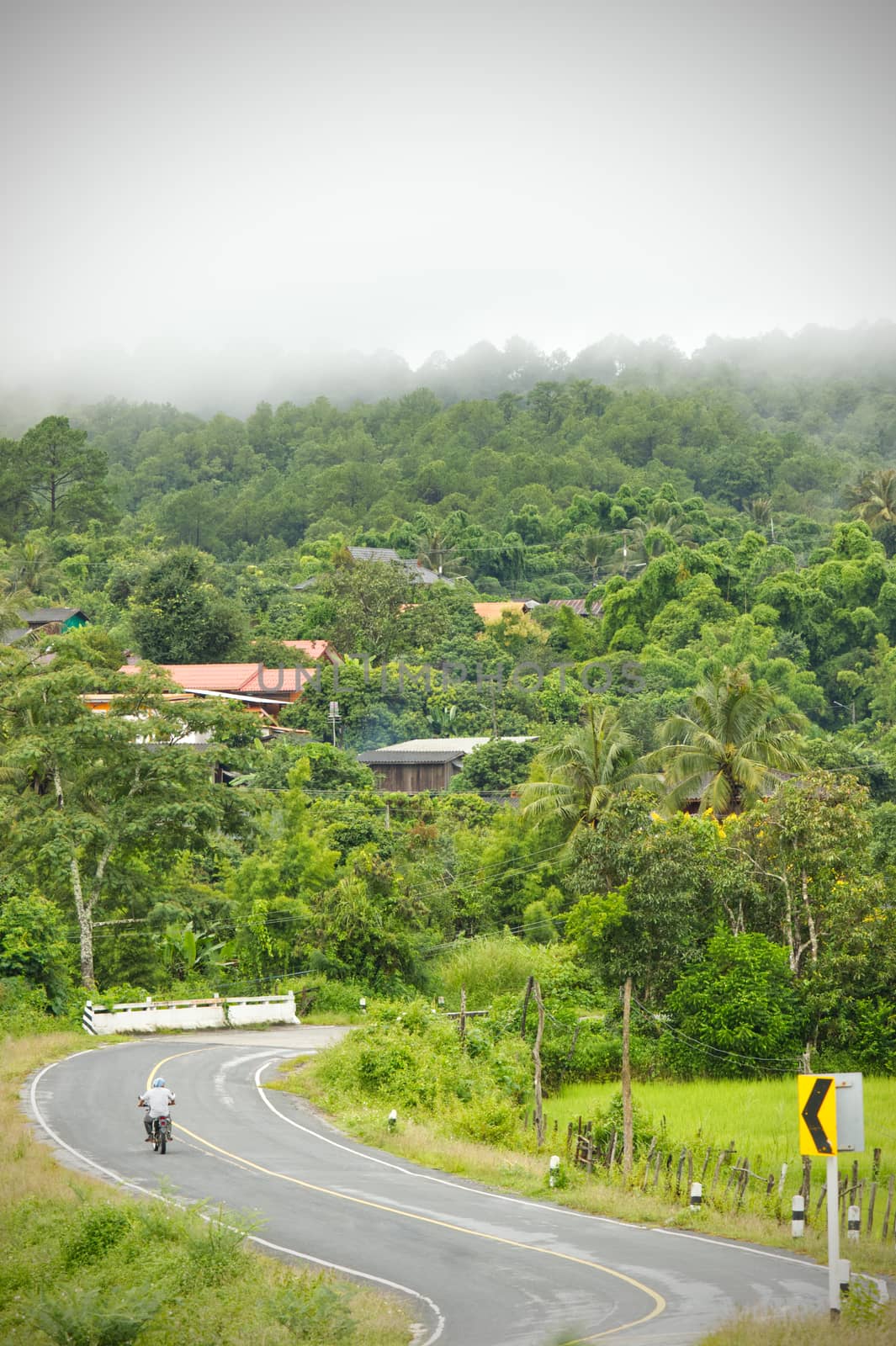 A motorcycle is running in the middle of a rural village road surrounded by mountains and fog in a refreshing morning. The concept of living in the nature of people is complete and balanced.