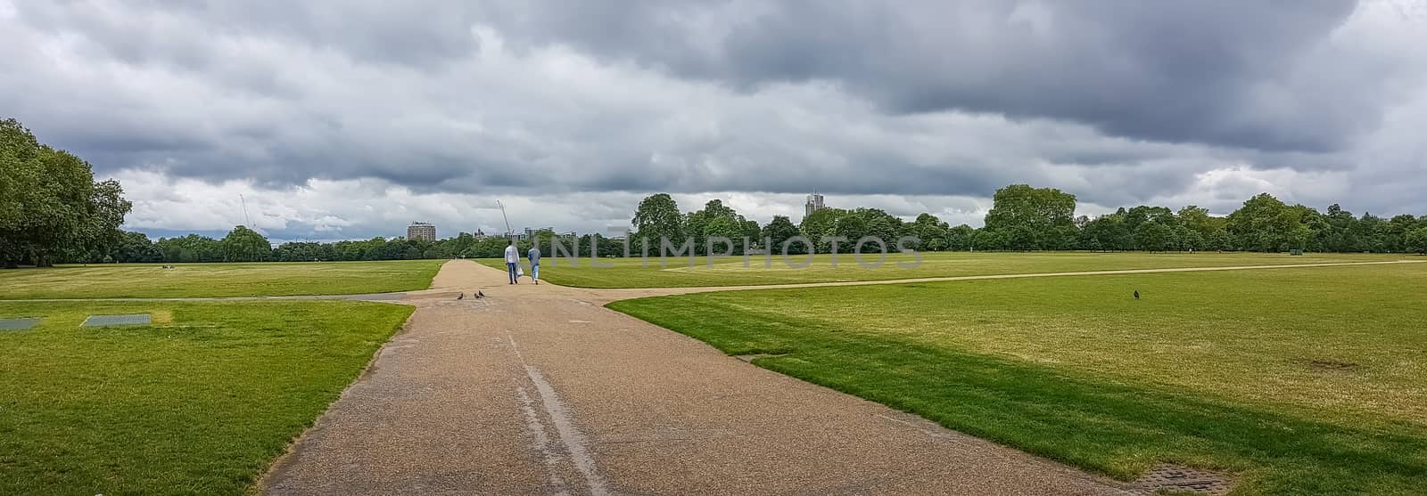 Panorama of Hyde Park field with two unrecognizable people walking in the middle of it. Cloudy sky as a background.