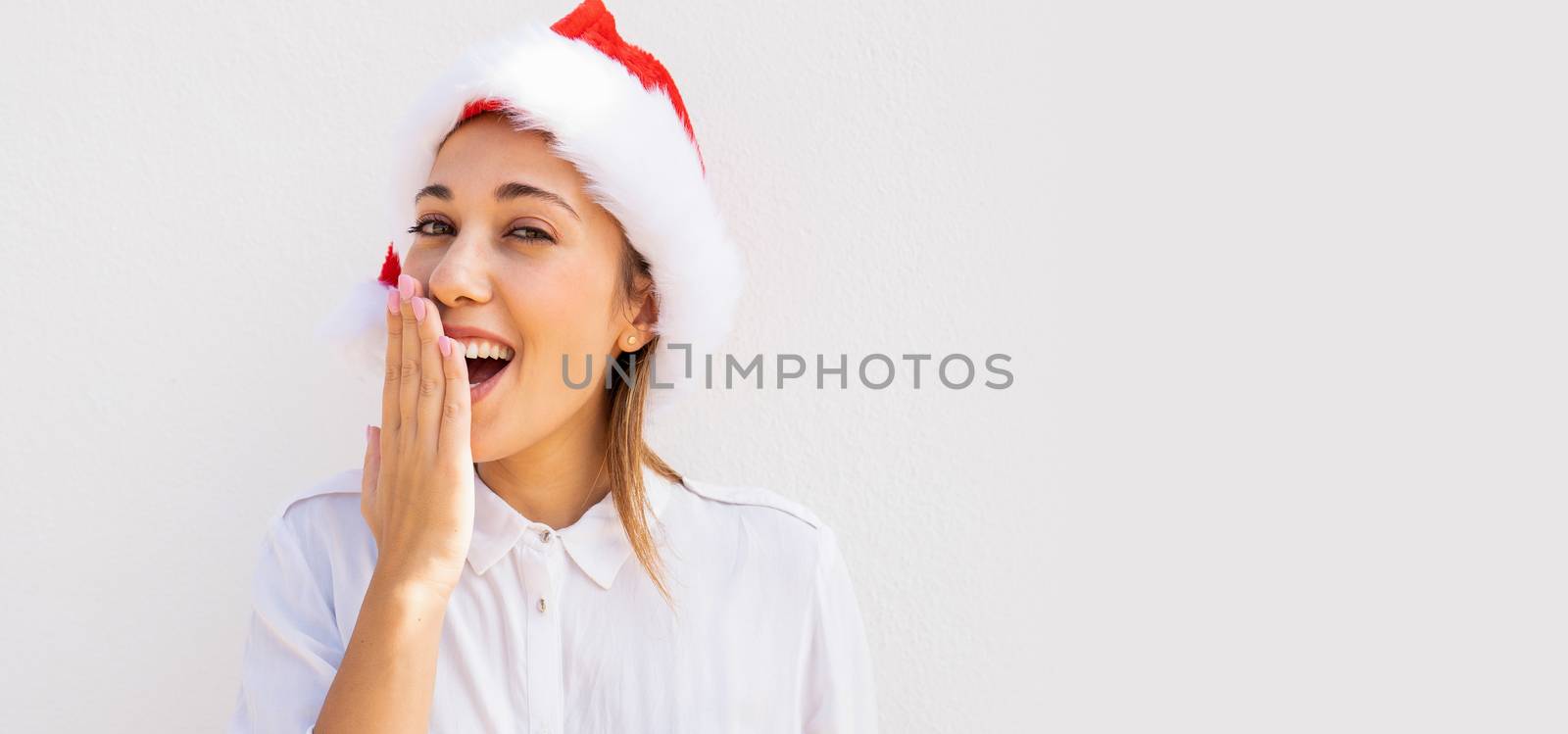 Young beautiful caucasian blonde woman with a Santa Claus hat surprising with his hand in front of his mouth with large white copy space background - Wow expression in Christmas time by robbyfontanesi